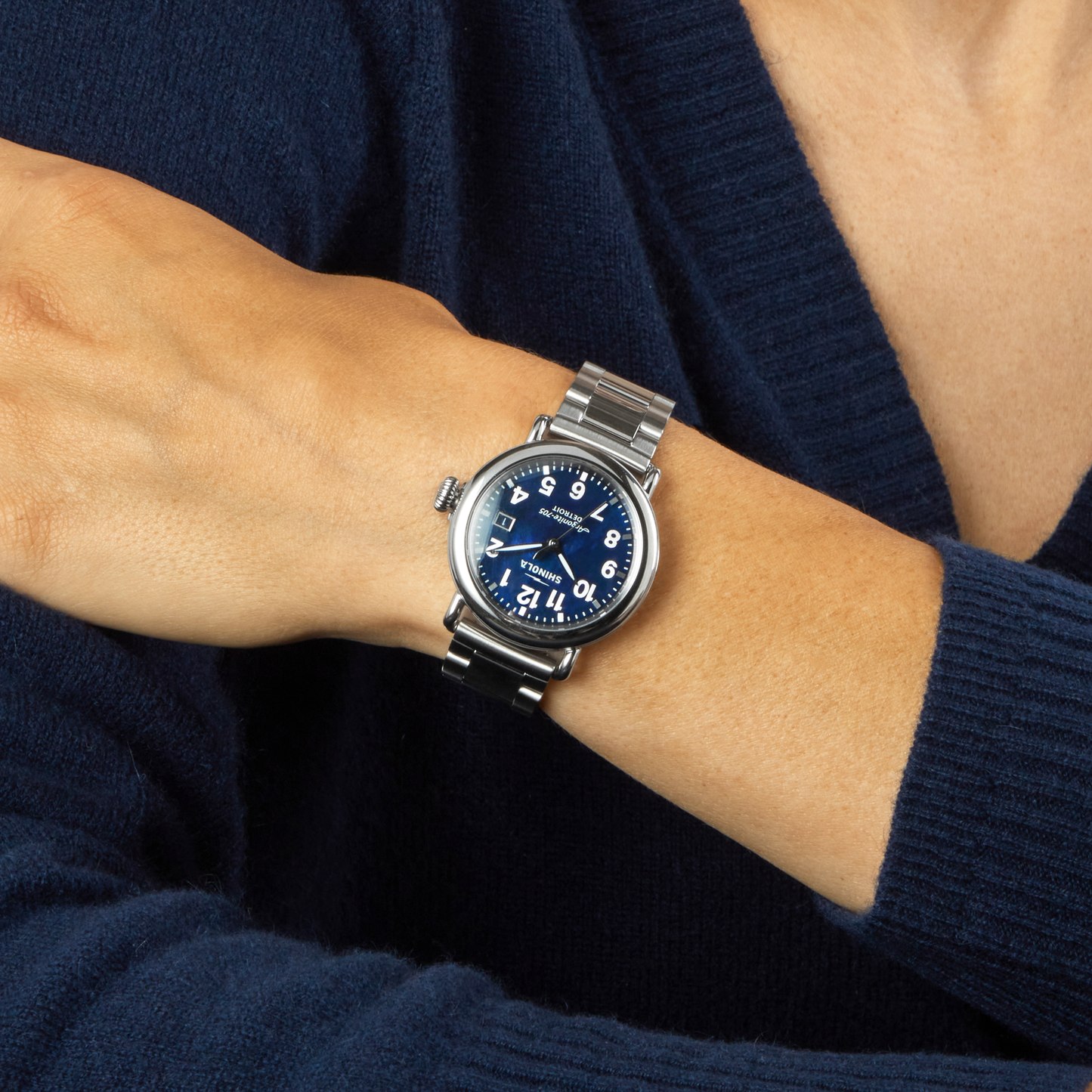 A person wearing the Shinola Runwell Watch - Cadet Blue, featuring a stainless-steel bracelet and blue face with white numbers, is dressed in a dark blue long-sleeve top. The watch appears on their left wrist.