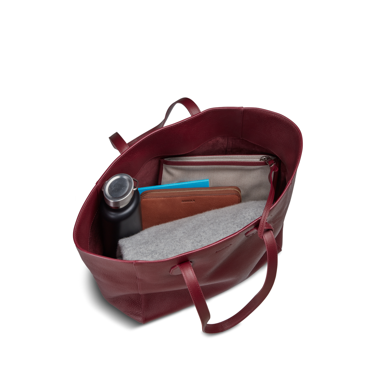A Shinola Runabout Tote Bag - Ruby, holding a water bottle, brown wallet, gray felt pouch, beige zipper pouch, and blue notebook, is viewed from above against a white background.