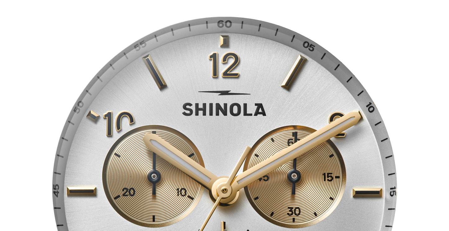 Close-up of the Shinola Canfield Sport Chronograph Watch - Silver, featuring gold hour markers and hands, subdials at 3 and 9 o’clock, a silver face, and the Shinola logo at center.