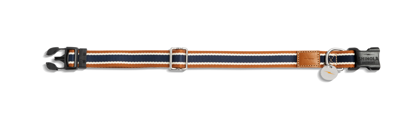 The Shinola Nylon Dog Collar - Brand Stripe features navy, white, and orange bands, a black plastic buckle, adjustable metal slider, leather patch accent, and two metal tags attached to a ring.