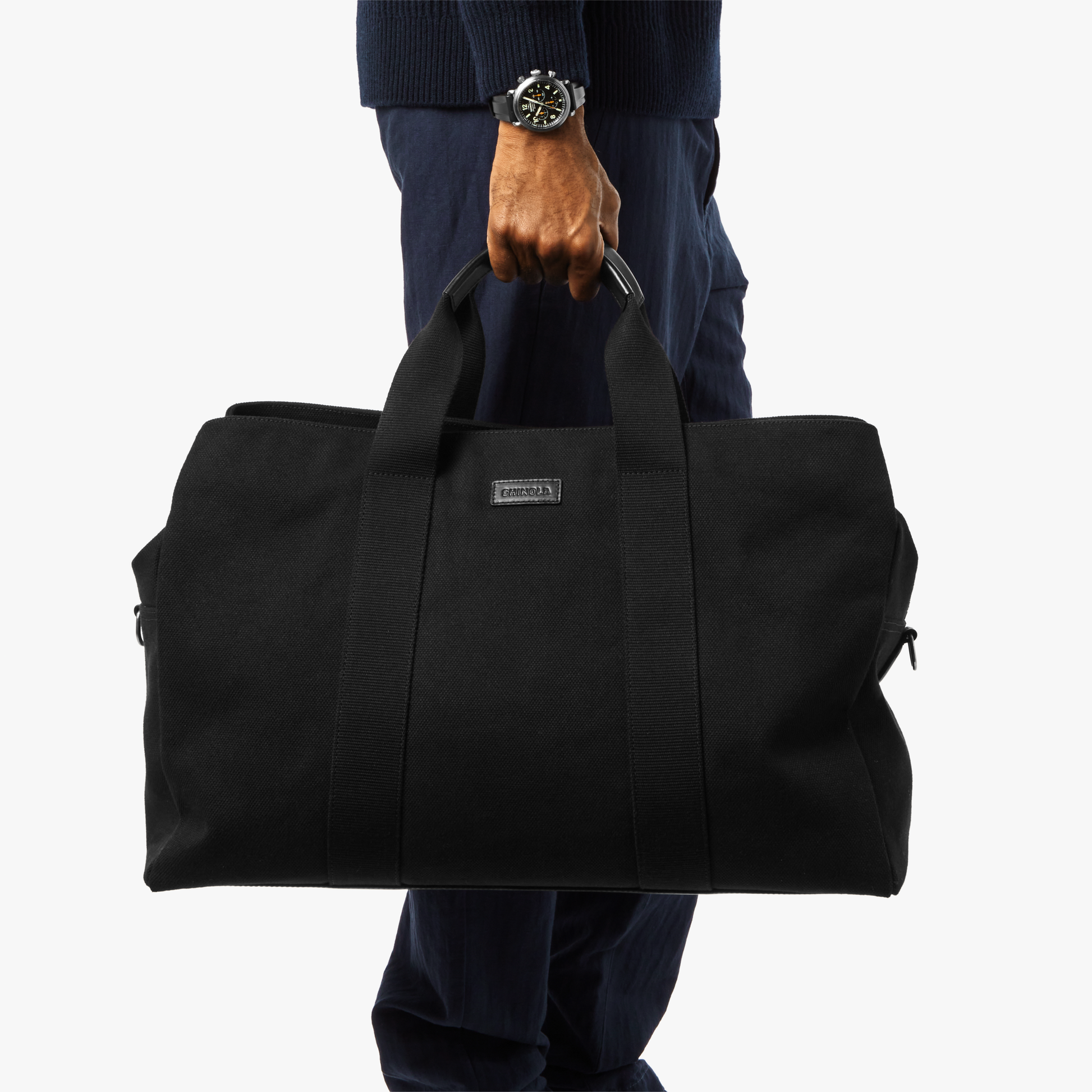 A person wearing a dark sweater, blue pants, and a watch holds the Shinola Runwell Canvas Duffle Bag - Black with two handles, perfect as a TSA carry-on, standing against a plain white background.