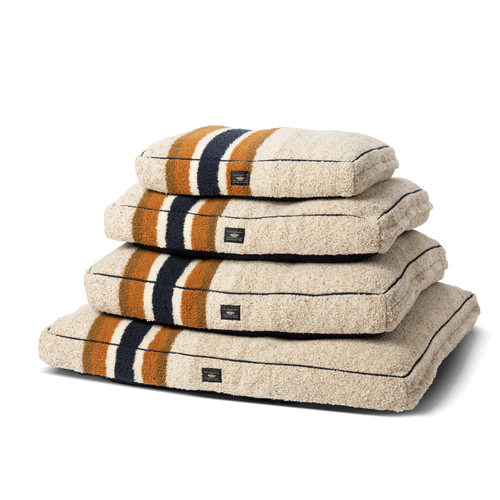 Four Napper Pillow Dog Beds by Shinola in beige with black, brown, and orange stripes are stacked together. Each features textured fabric, a small black front label, and comfy memory fiber fill.