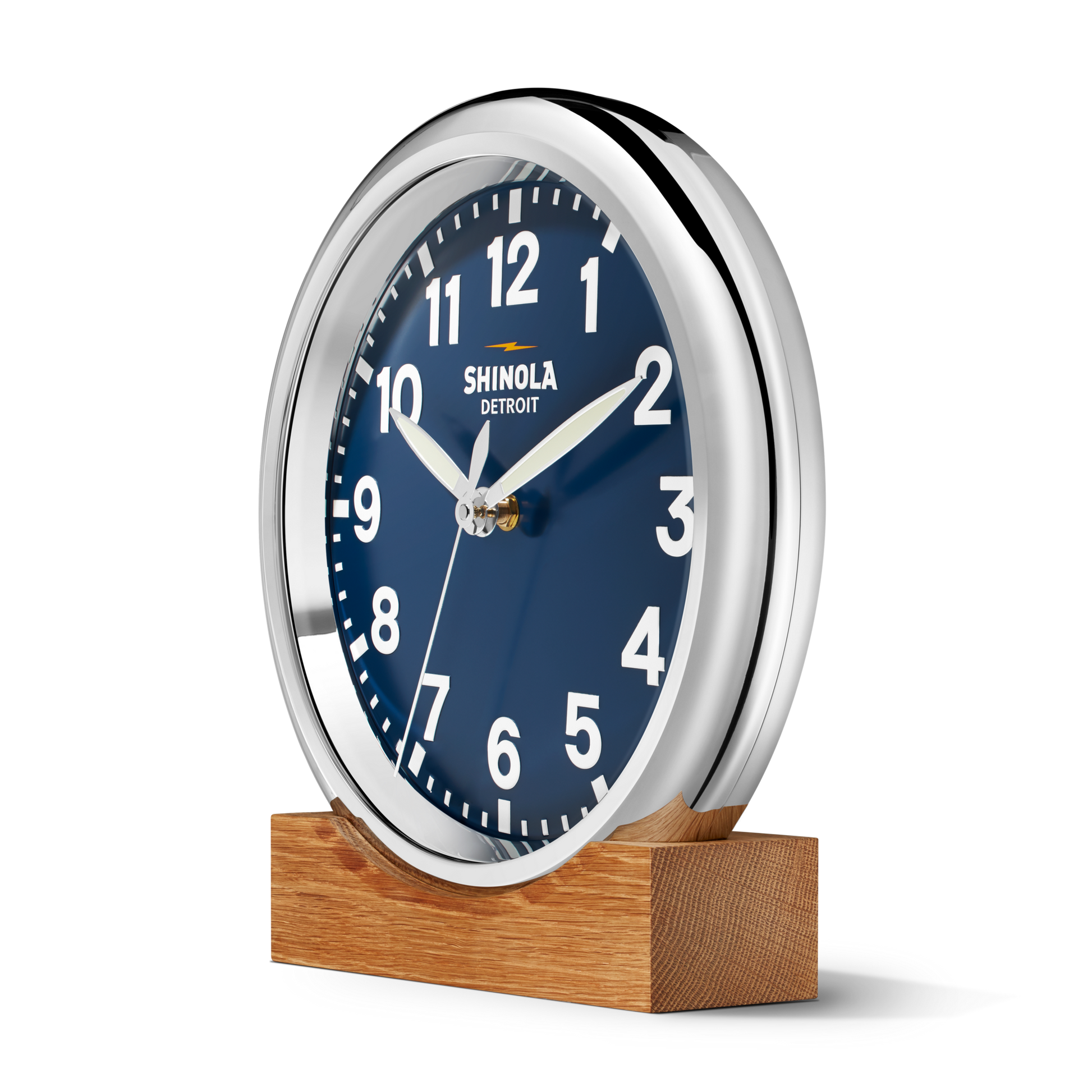 A 14" Shinola Runwell Clock - Navy with a matte midnight blue dial, white numbers and shiny silver frame sits on a rectangular wooden base. The face reads 10:10 and displays "Shinola Detroit.