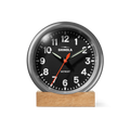 A Shinola 6" Runwell Clock in tumbled zinc and black, featuring white numbers, a red second hand, and silver hands, rests on a rectangular wooden base against a plain white background.