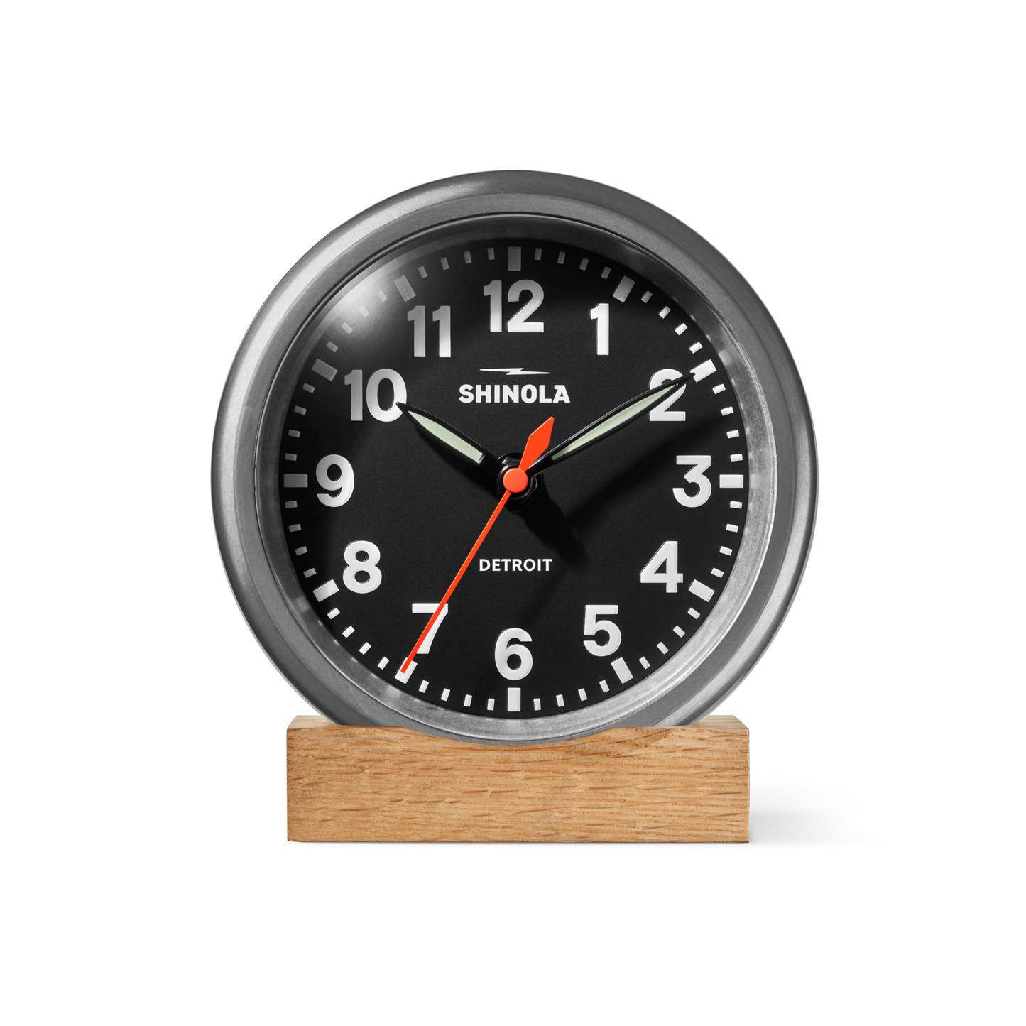A Shinola 6" Runwell Clock in tumbled zinc and black, featuring white numbers, a red second hand, and silver hands, rests on a rectangular wooden base against a plain white background.