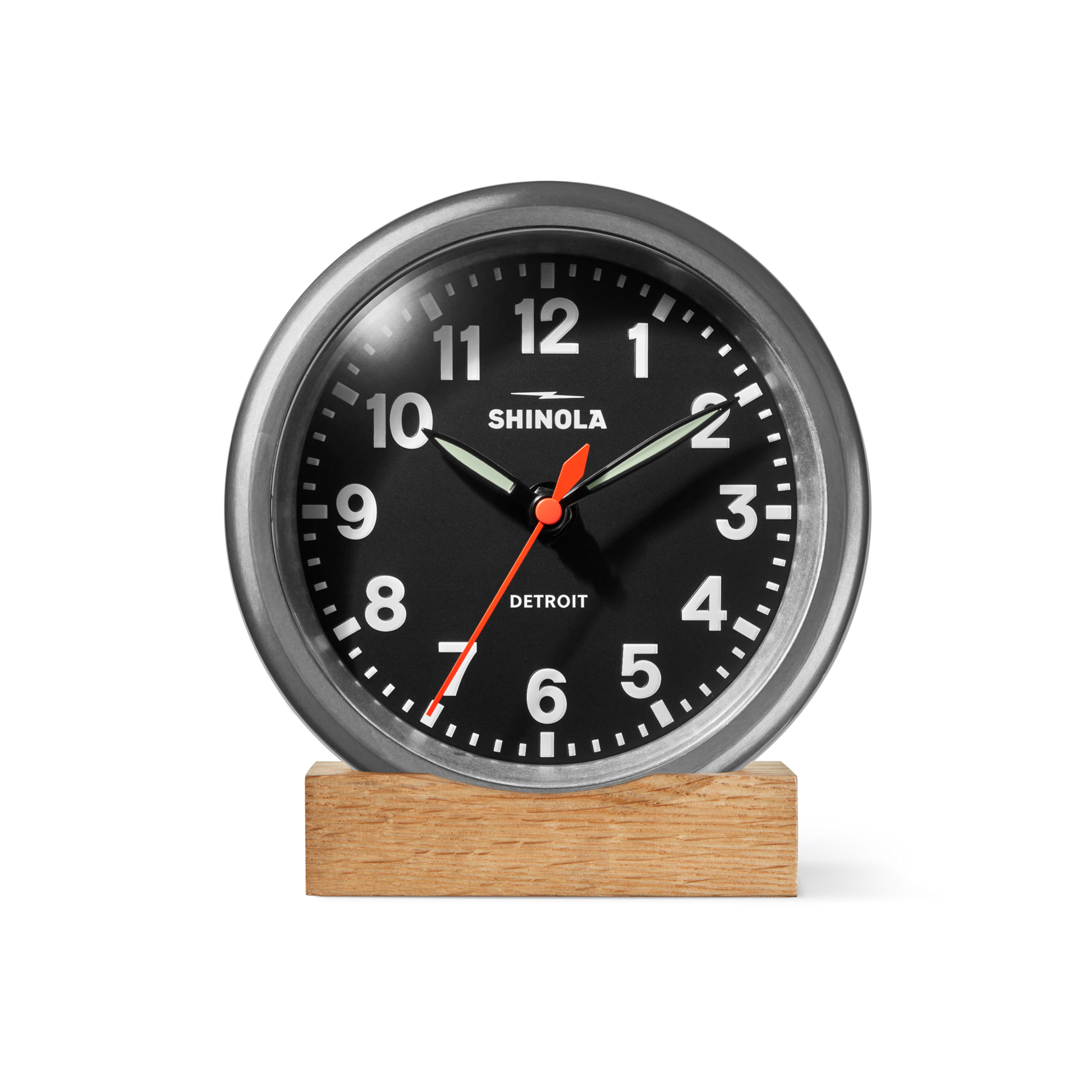 A Shinola 6" Runwell Clock in tumbled zinc and black, featuring white numbers, a red second hand, and silver hands, rests on a rectangular wooden base against a plain white background.