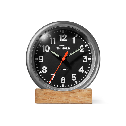 A Shinola 6" Runwell Clock in tumbled zinc and black, featuring white numbers, a red second hand, and silver hands, rests on a rectangular wooden base against a plain white background.