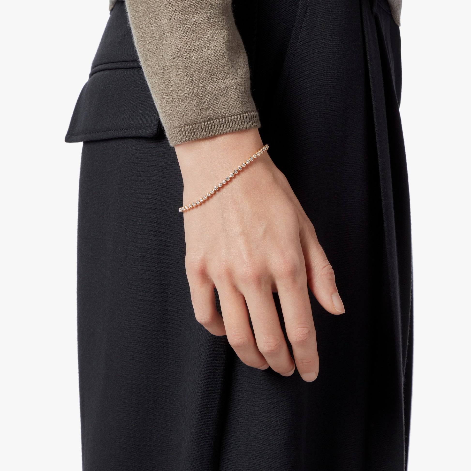 A close-up shows a person's left hand and wrist wearing the Shinola 1 Carat 7" Diamond Tennis Bracelet in yellow gold. The person, dressed in a taupe long-sleeve top and black pants, is set against a plain light background.