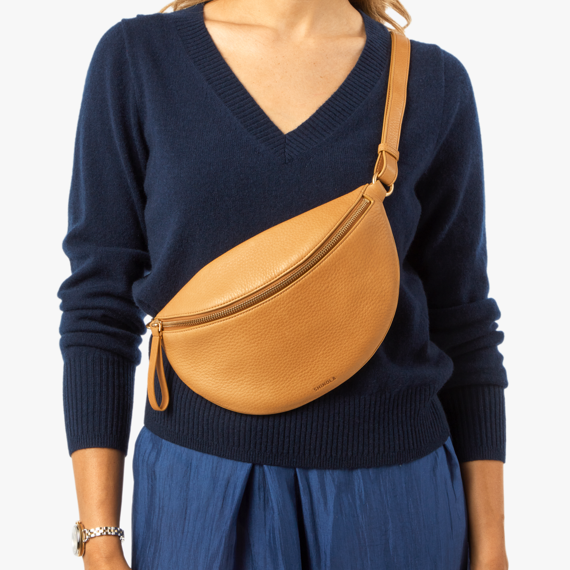 A woman in a navy blue sweater and blue skirt carries the Shinola Sling Bag - Honey, a tan leather crossbody with an adjustable strap and zipper, slung diagonally from her shoulder to waist.