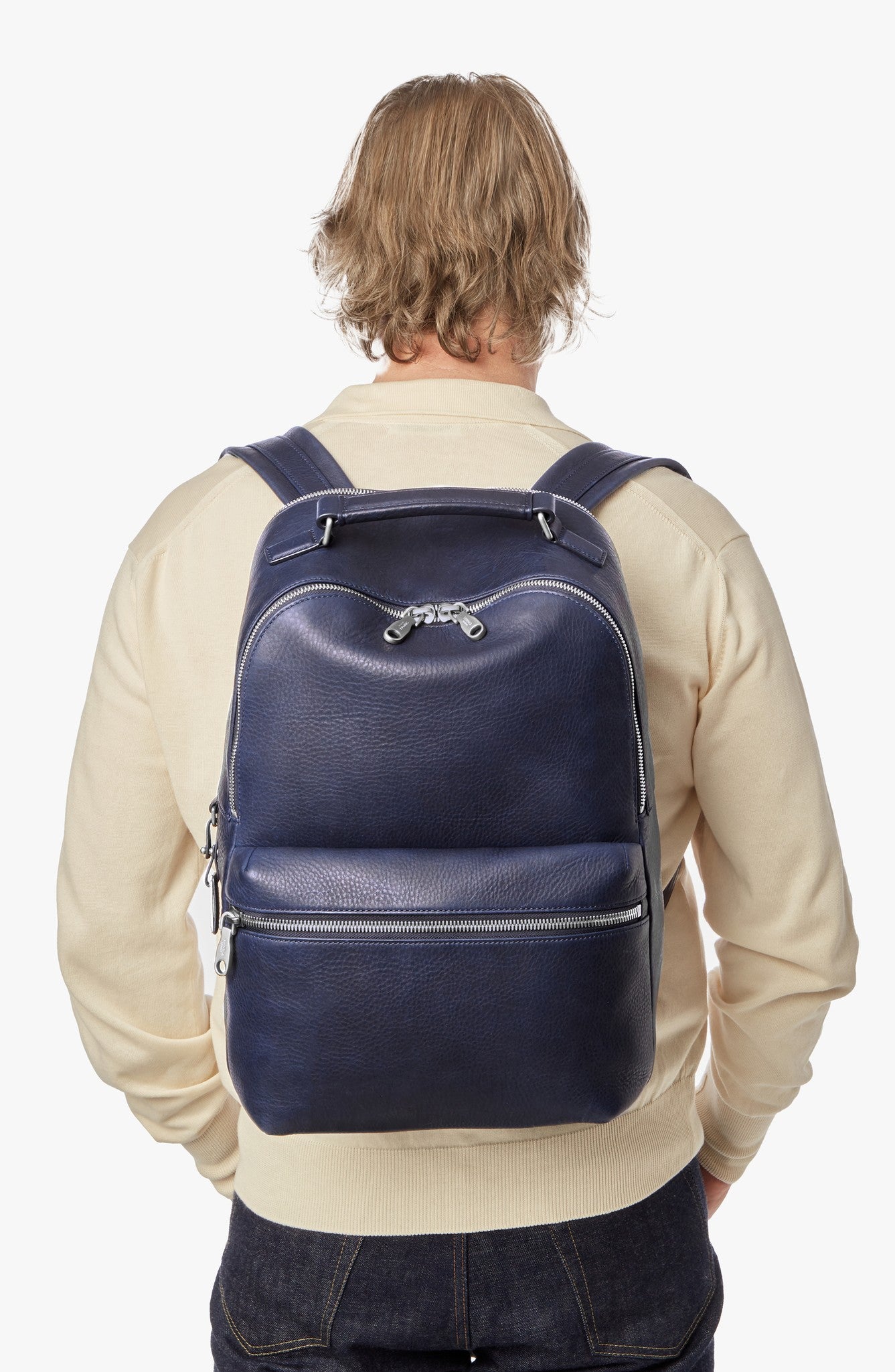 A person with blonde hair, wearing a cream sweater and dark jeans, is shown from behind carrying a large navy leather backpack and holding the Shinola Portfolio Briefcase in Stone.