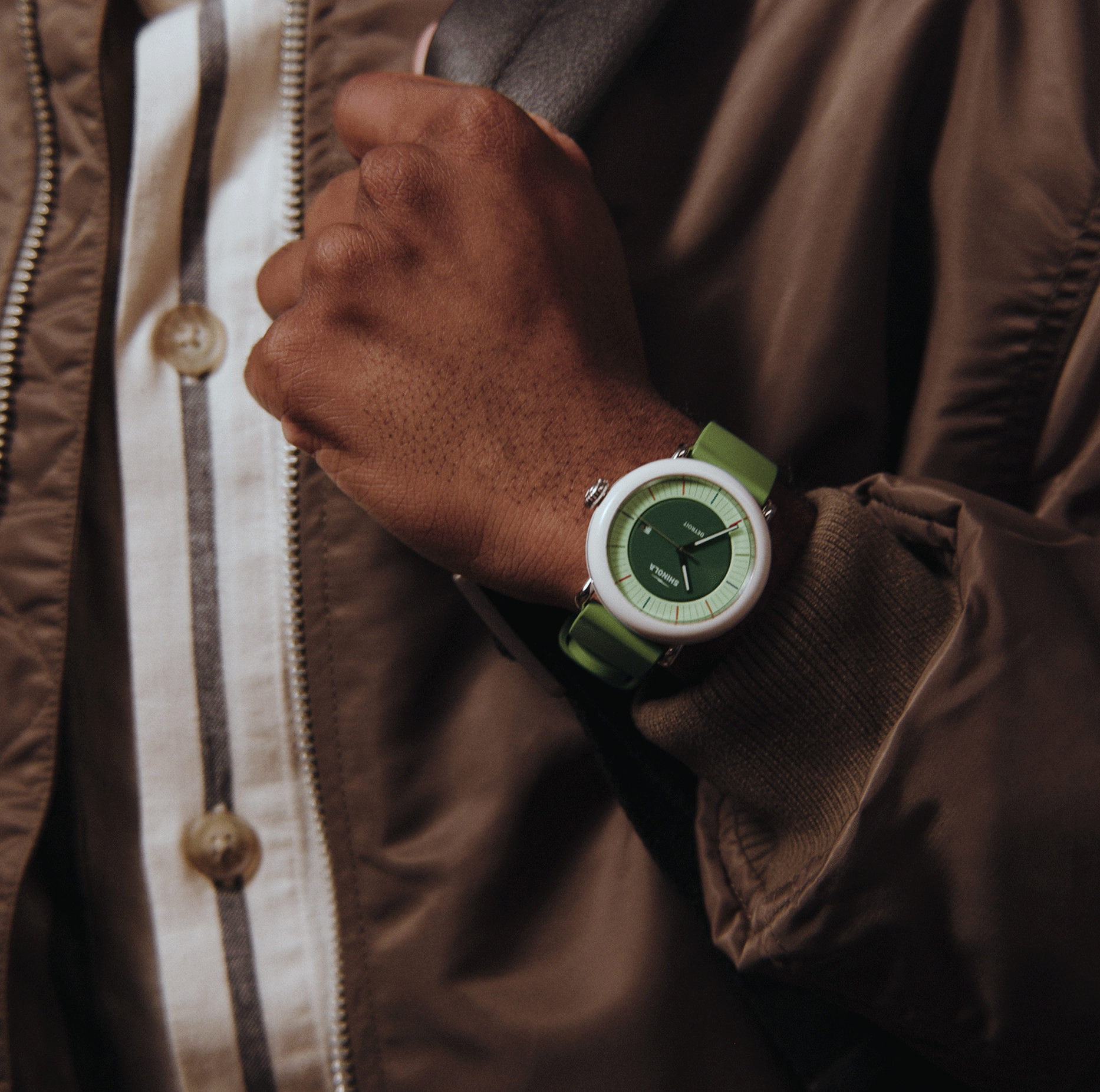 A person in a brown jacket and striped shirt holds a strap over their shoulder, wearing a Shinola Detrola Watch - Deep Olive with a white face, green strap, and silver details on their left wrist.