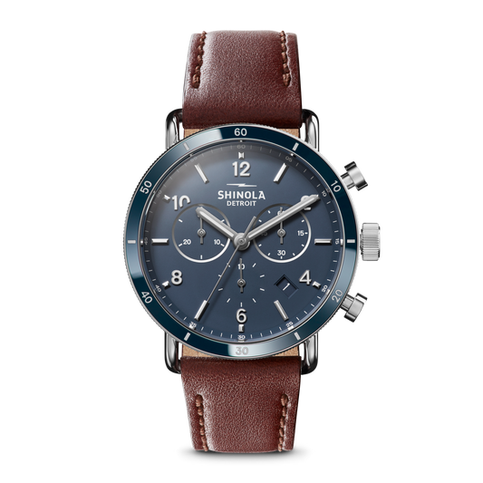 The Shinola Canfield Sport Chronograph Watch - Midnight Blue features a blue dial with silver hands and markings, three subdials, a chronograph function, and a brown leather strap. The watch face reads Shinola Detroit on a plain white background.