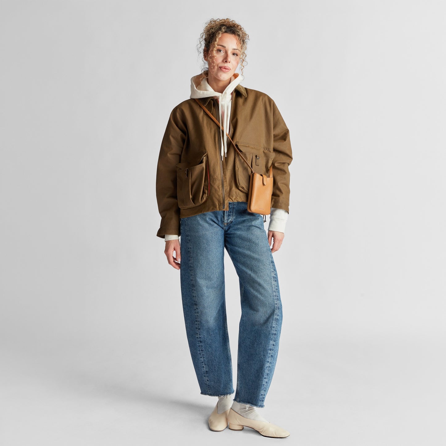 A person with curly hair stands against a plain background wearing a brown jacket, white hoodie, blue jeans, beige shoes, and holding the Shinola Bixby Phone Crossbody in Tan—ideal for traveling light.
