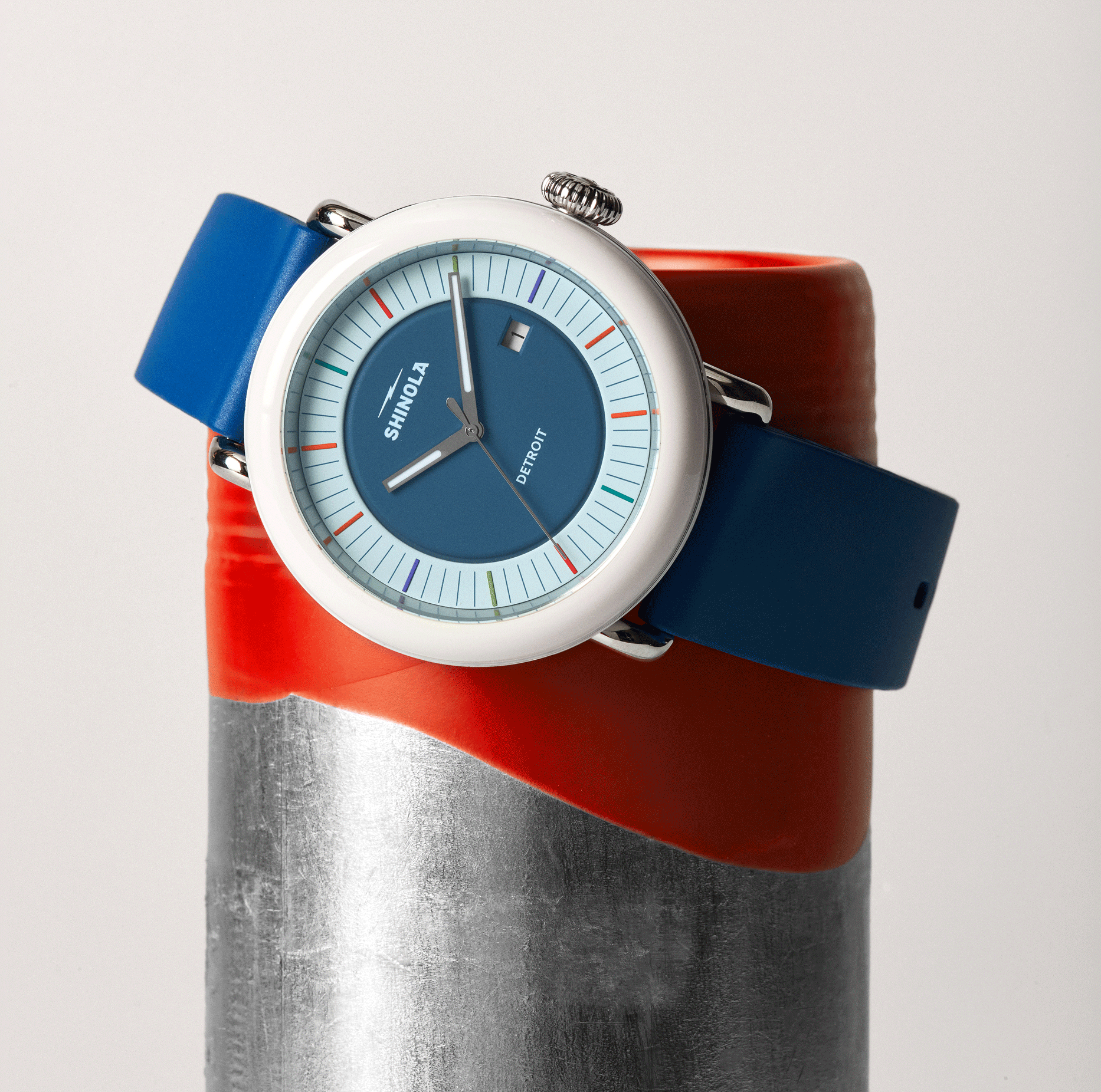 The Shinola Detrola Watch - Blue, featuring a round face and white bezel, is showcased on a cylinder with a silver base and red top against a light background—an eye-catching, modern timepiece.