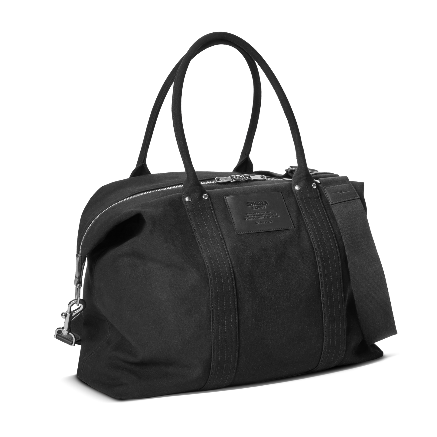 The Shinola Runabout Holdall Canvas Bag in black features double handles, a detachable shoulder strap for crossbody wear, silver zipper, and a rectangular stitched front patch. Its structured canvas design offers a minimalist look.