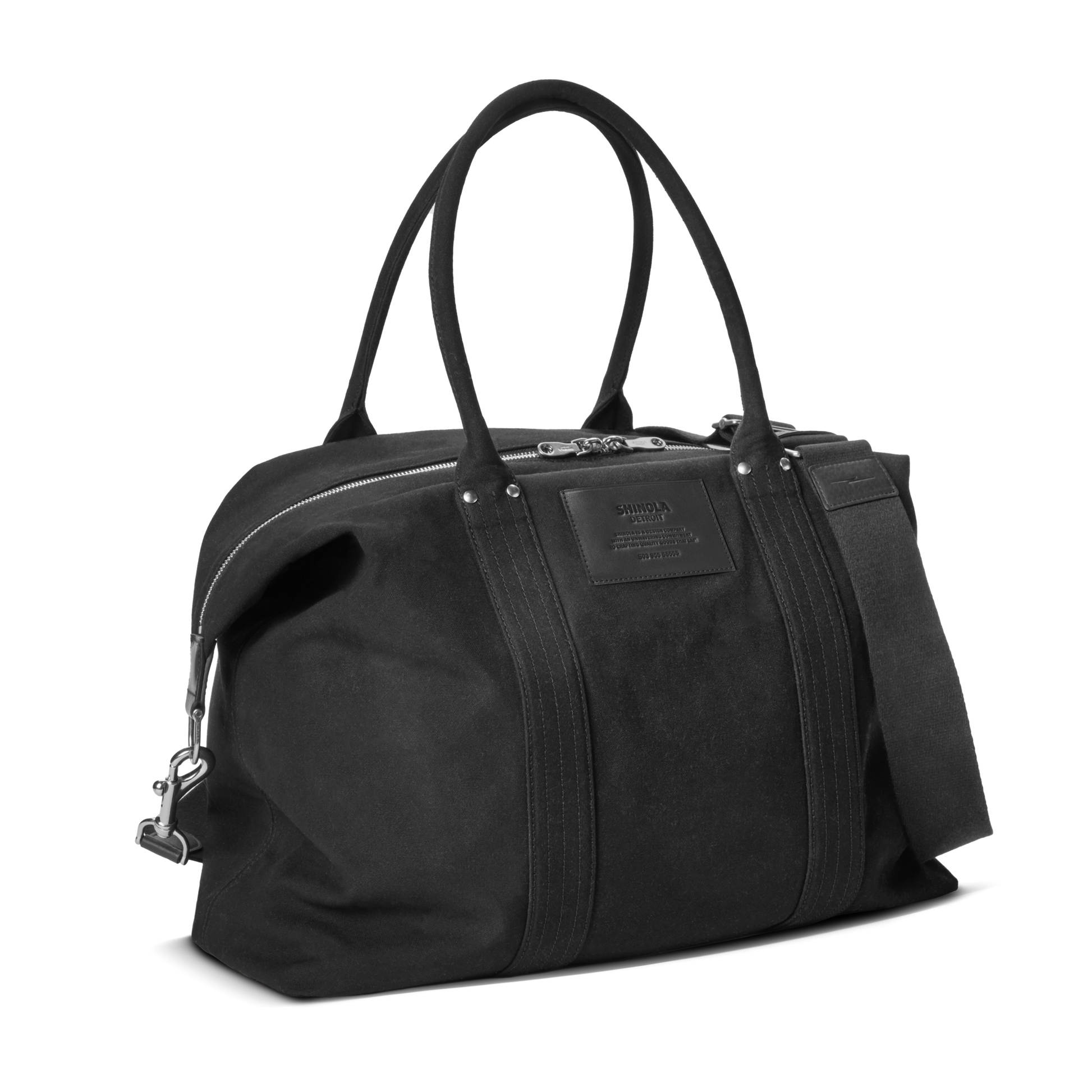 The Shinola Runabout Holdall Canvas Bag in black features double handles, a detachable shoulder strap for crossbody wear, silver zipper, and a rectangular stitched front patch. Its structured canvas design offers a minimalist look.