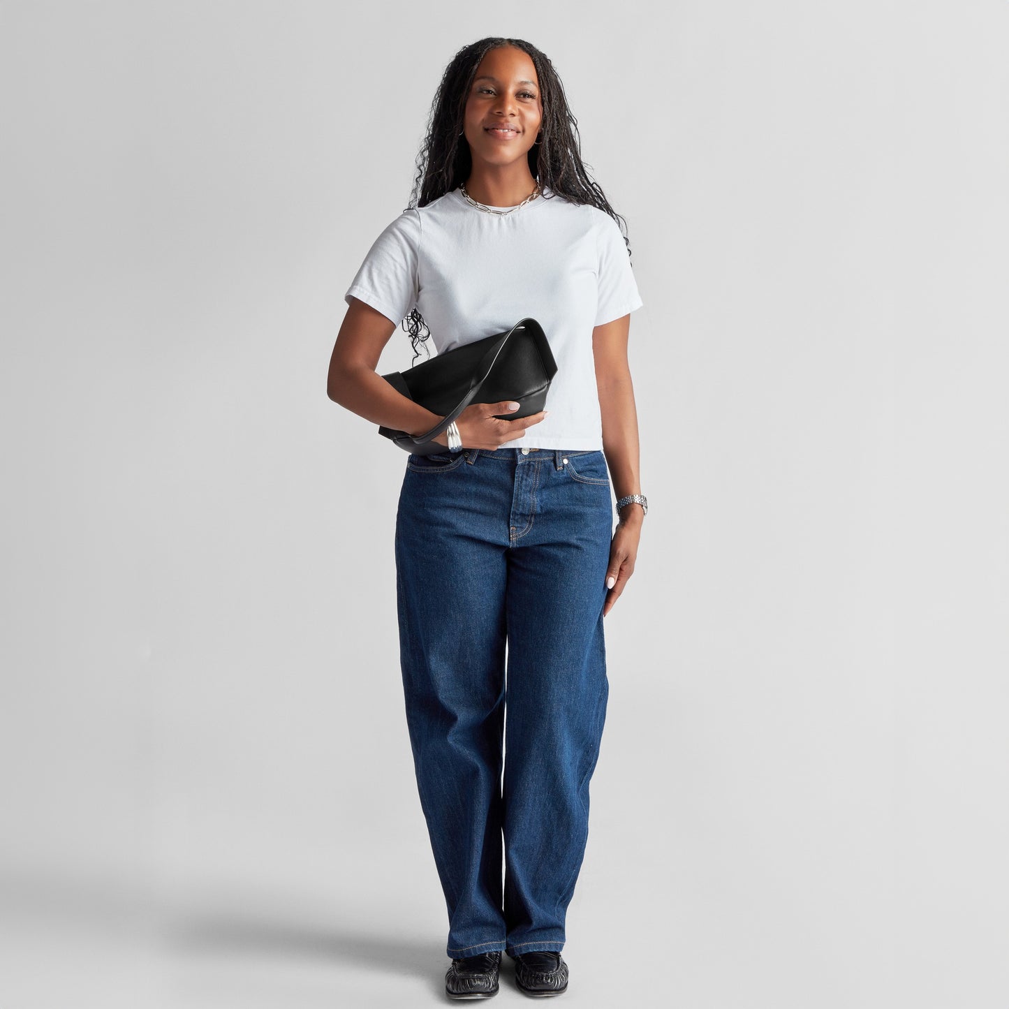 A woman stands against a plain background, smiling. She is wearing a white t-shirt, blue jeans, black shoes, and holds a Shinola Small Derby Shoulder Bag in black under her arm—a chic accessory for day or night.