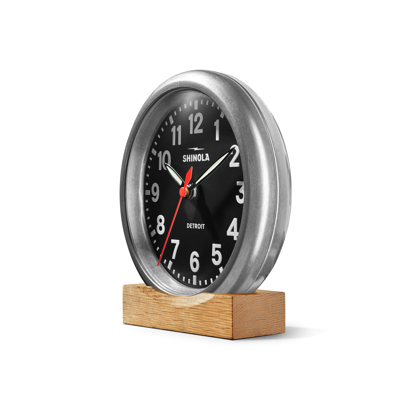 A Shinola 6" Runwell Clock in tumbled zinc with a black face, white numbers, and a red second hand sits on a rectangular wooden base, reading 10:10 and displaying the Shinola brand name.
