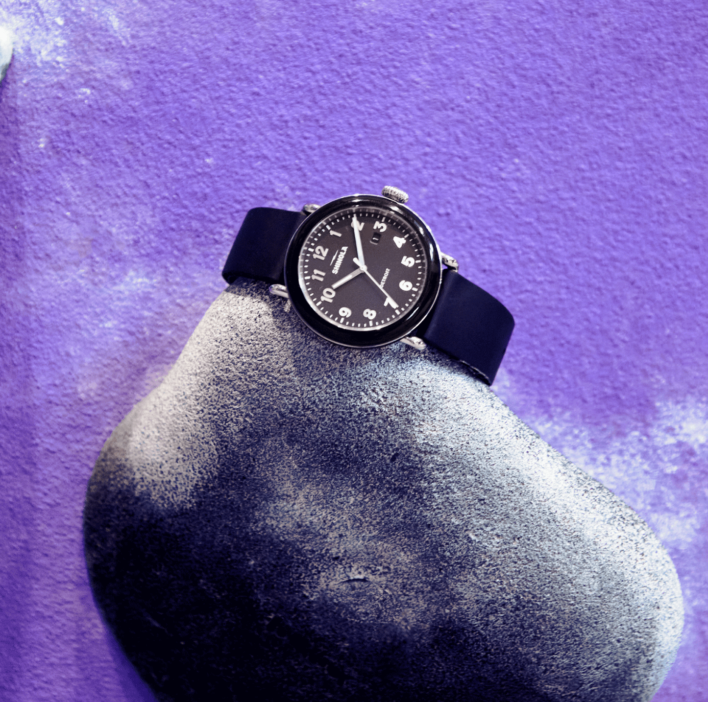 A Shinola Detrola Watch - Black with a round face and white numbers rests on a gray, textured rock against a purple background, highlighting classic American design.