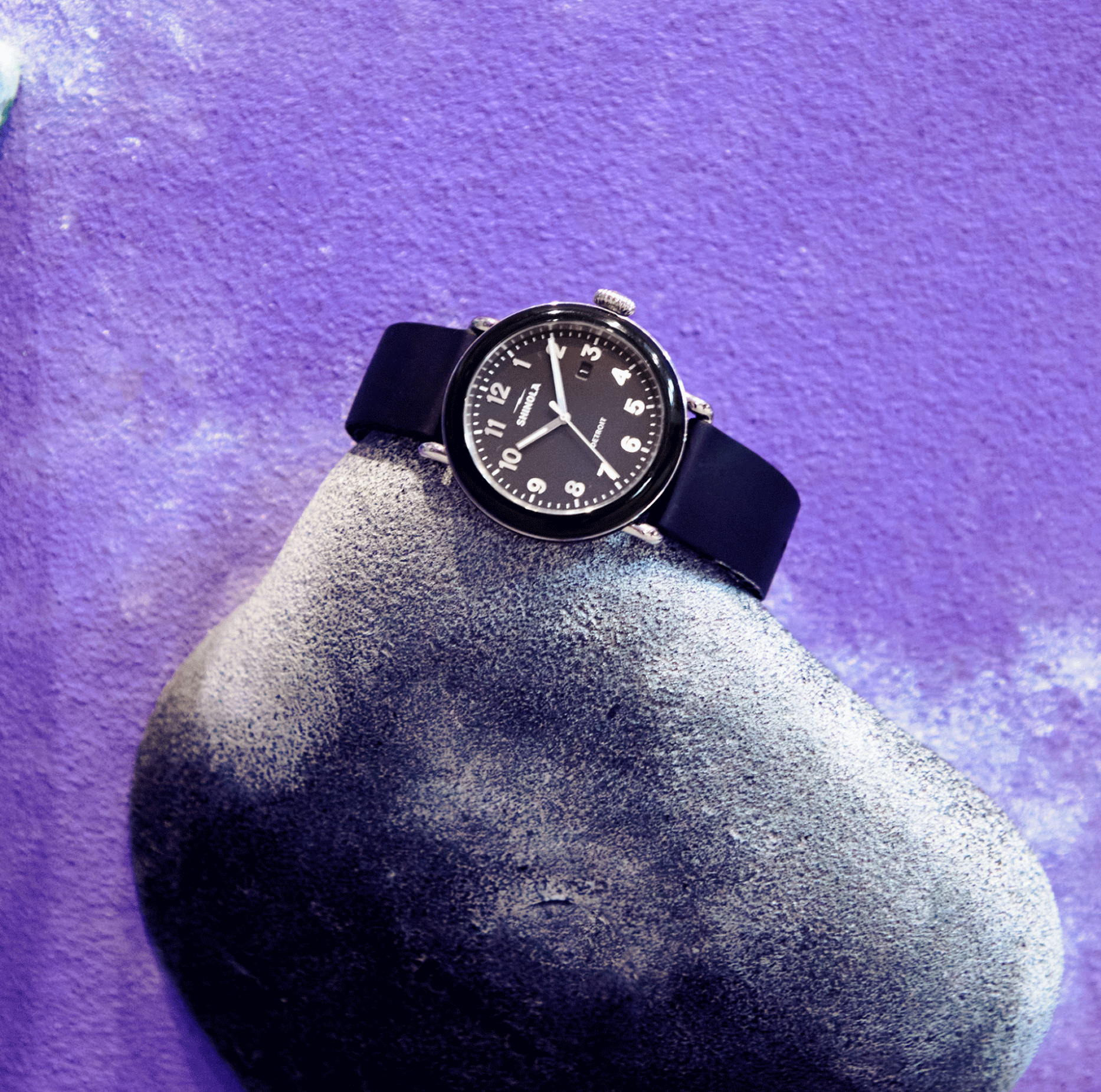 A Shinola Detrola Watch - Black with a round face and white numbers rests on a gray, textured rock against a purple background, highlighting classic American design.