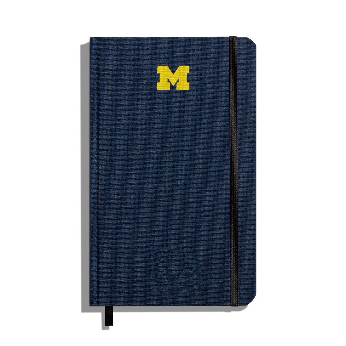 The Shinola Medium Hard Ruled Journal in navy features a yellow University of Michigan M logo on the cover, a black elastic band closure on the right side, and a black ribbon bookmark at the bottom.
