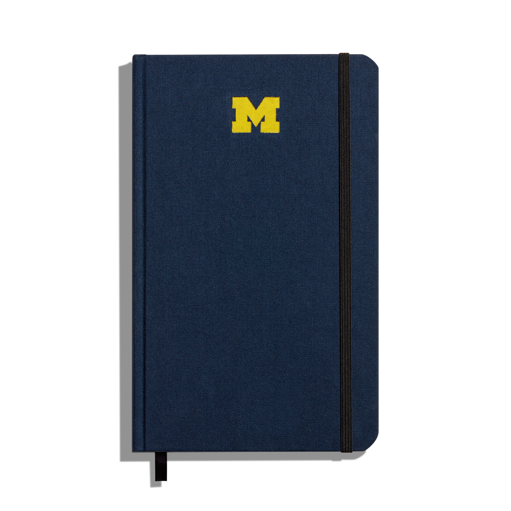 The Shinola Medium Hard Ruled Journal in navy features a yellow University of Michigan M logo on the cover, a black elastic band closure on the right side, and a black ribbon bookmark at the bottom.