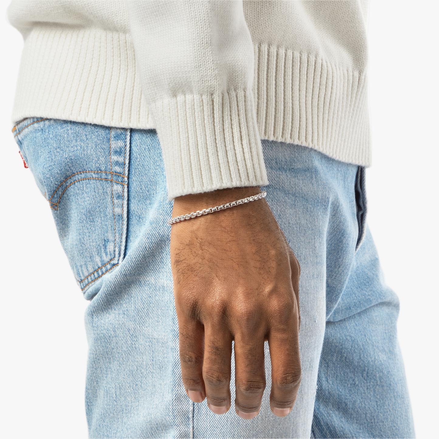 A person in a white sweater and light blue jeans stands with their hand by their side, wearing the Shinola 4mm Box Chain Bracelet, a sterling silver accessory showcased on their wrist.