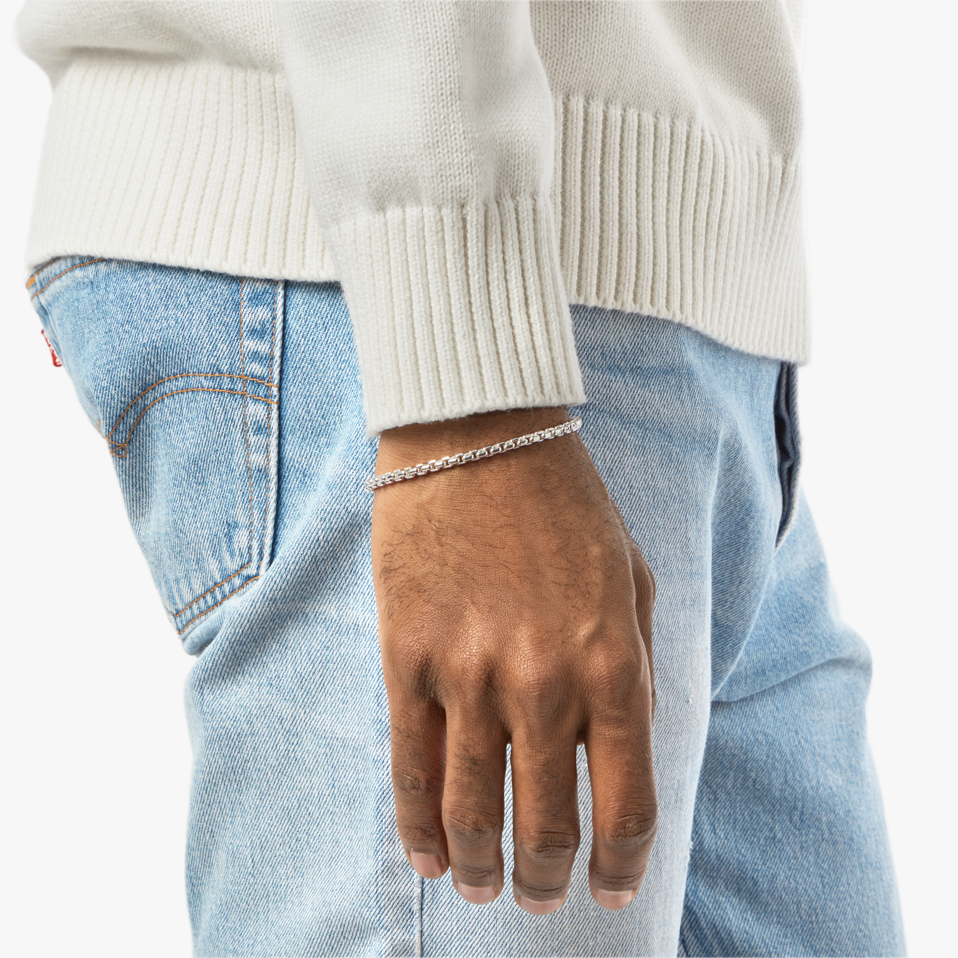 A person in a white sweater and light blue jeans stands with their hand by their side, wearing the Shinola 4mm Box Chain Bracelet, a sterling silver accessory showcased on their wrist.