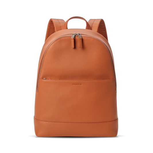 The Shinola Canfield Backpack in Rust is a sleek orange leather bag with a front zip pocket, dual zipper pulls on the main compartment, and padded shoulder straps—perfect for stylish travel—shown against a plain white background.