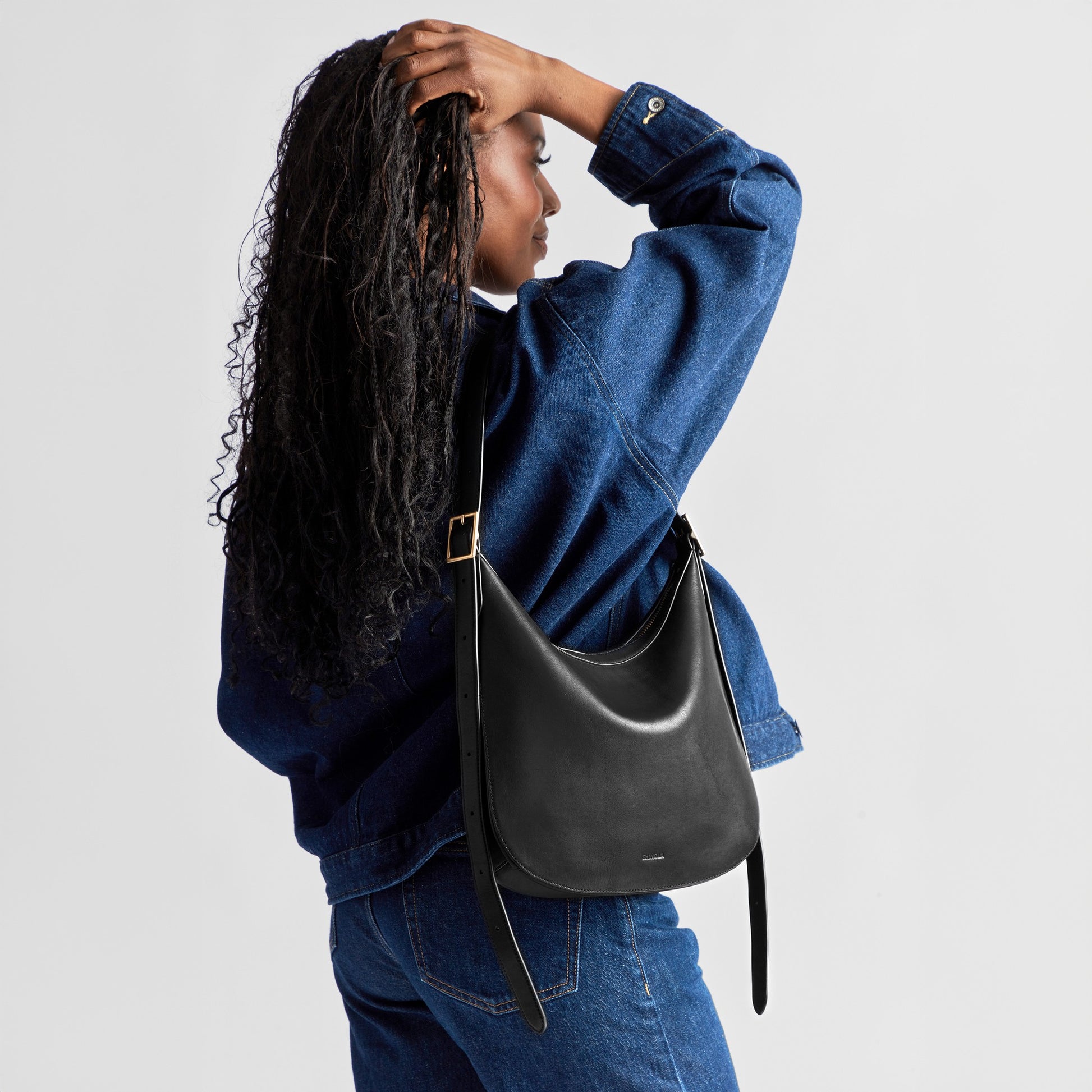 A woman with long curly hair in a blue denim jacket and jeans stands sideways, holding up her hair and carrying the Shinola Medium Bixby Crossbody - Black. The background is plain white.