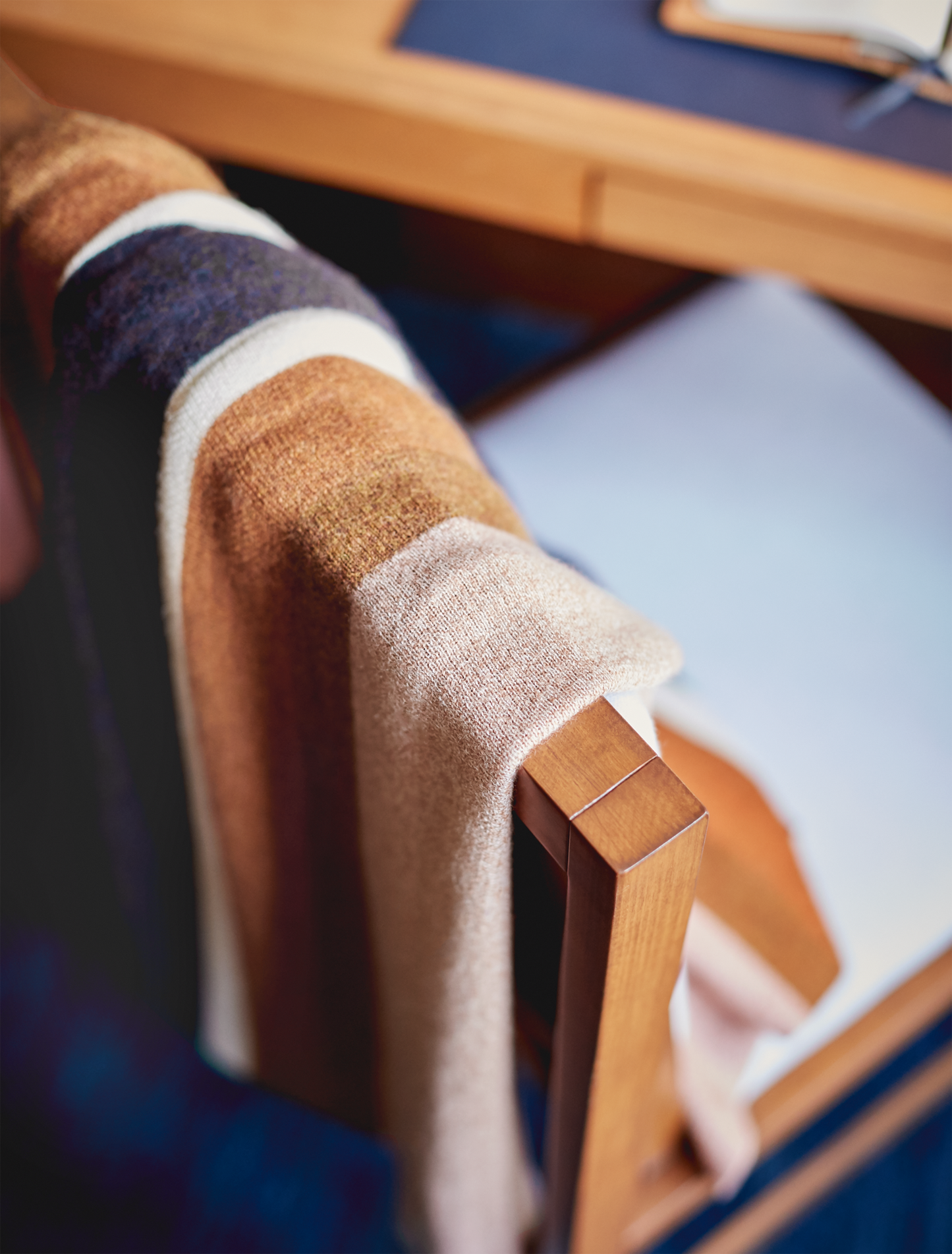 A soft, striped Shinola Alpaca Throw Blanket in pink and beige is draped over the back of a wooden chair beside a wooden desk with a notebook on top.