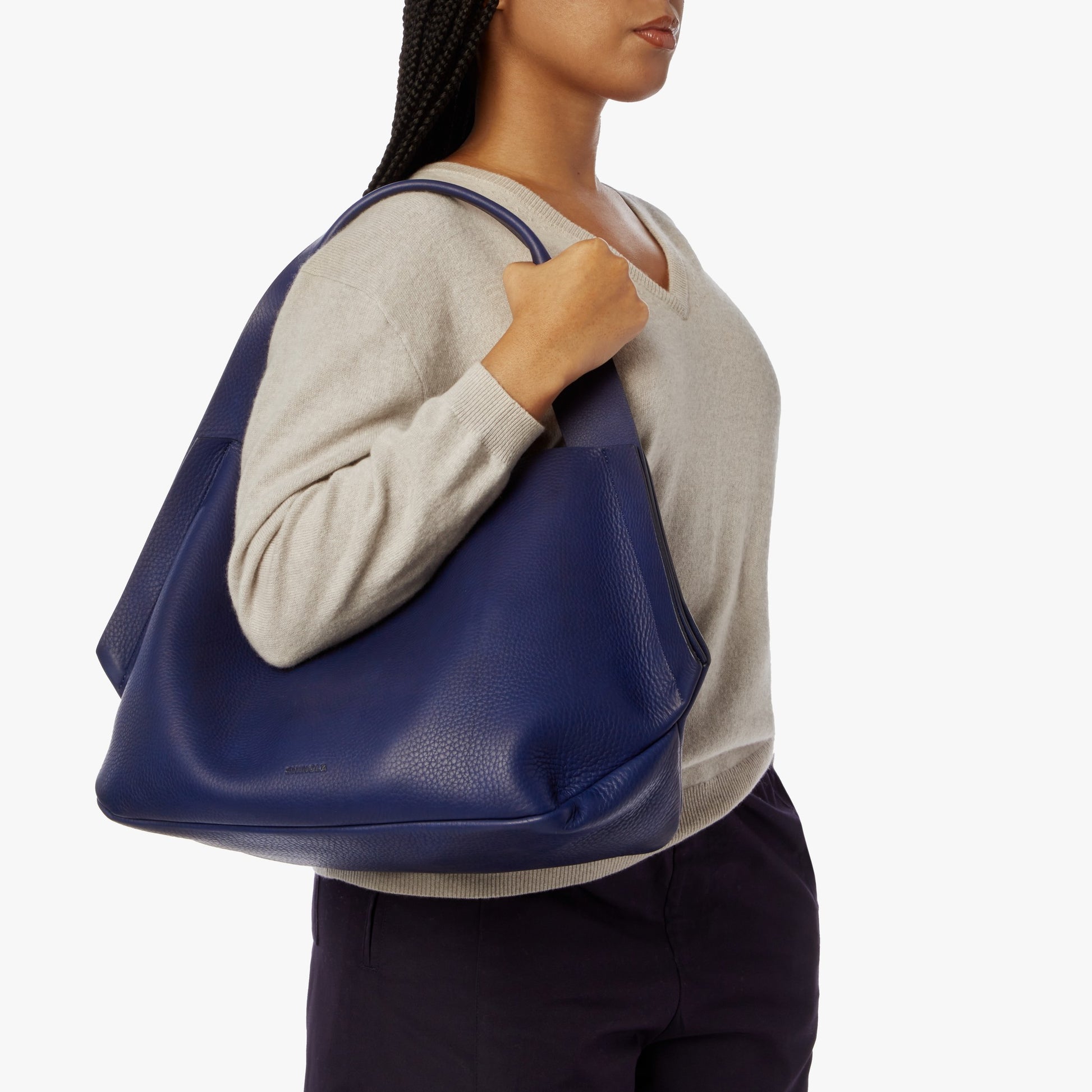 A woman in a light beige sweater and dark pants carries the Shinola Runwell Shoulder Bag - Lake, featuring stylish handles and deep blue leather, on her right shoulder against a plain white background.