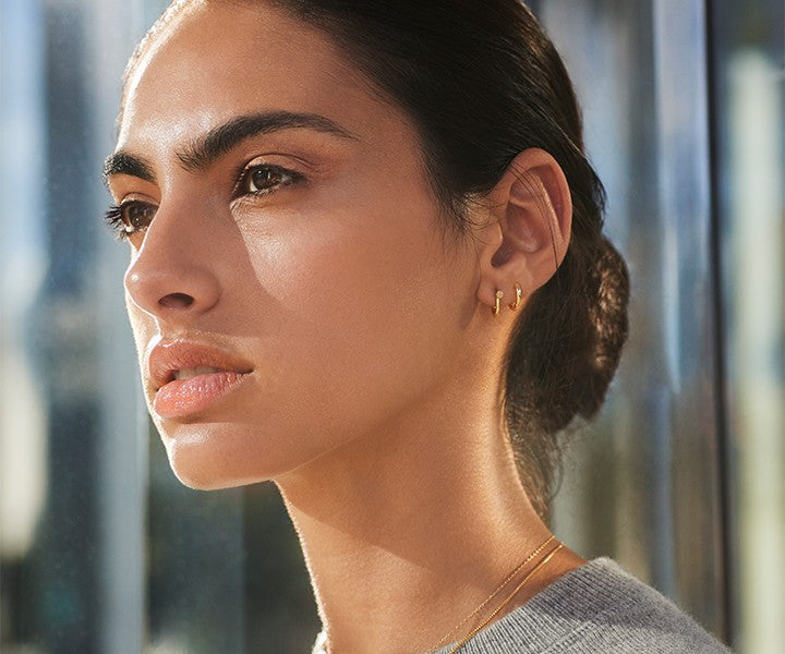 A woman with dark hair pulled back gazes thoughtfully, wearing Shinola’s Bike Lock Huggie Earrings in yellow gold and a delicate necklace, sunlight illuminating her face—an ideal everyday earring choice.