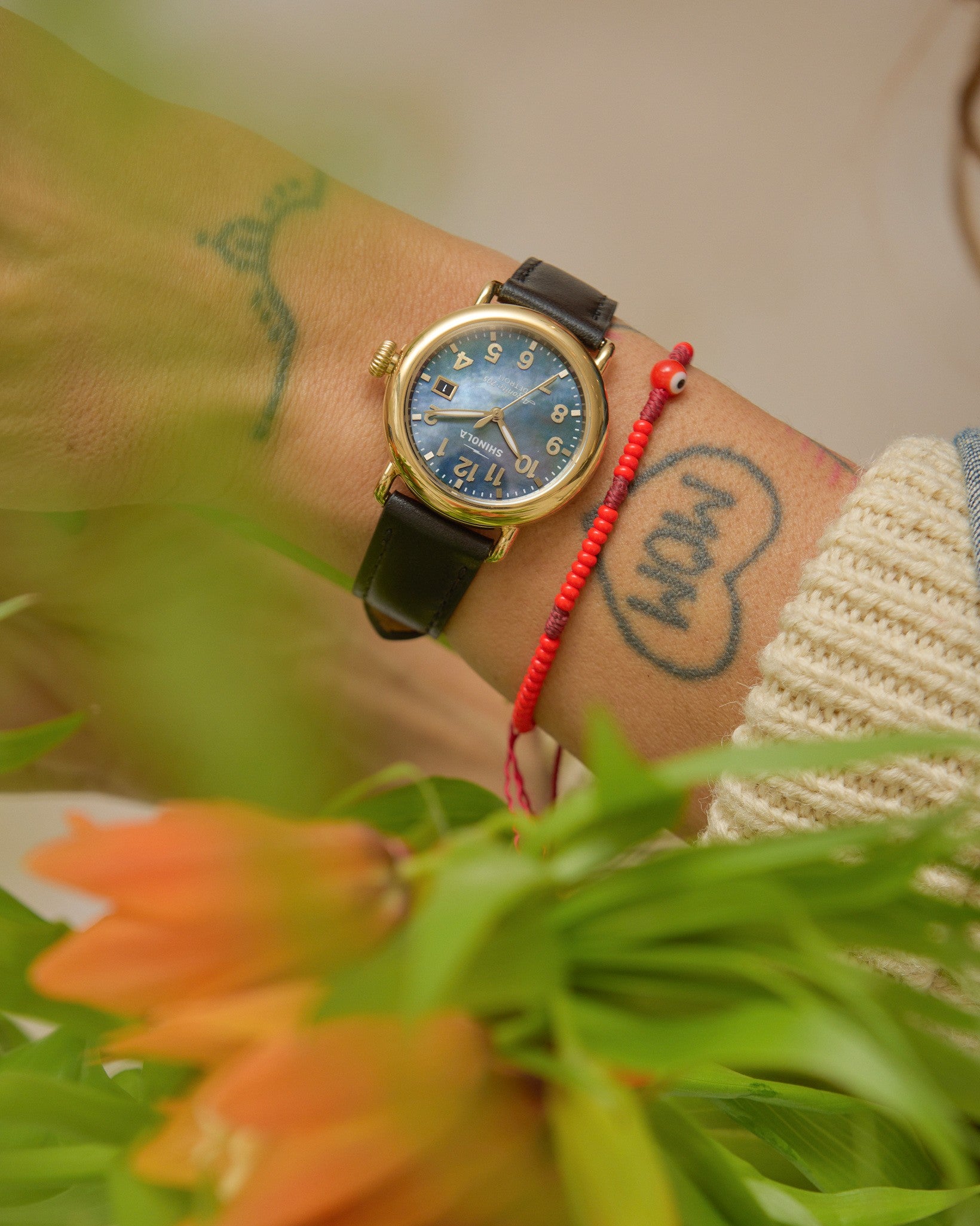 A close-up shows a wrist with a Shinola Runwell Watch—MOP Black Leather Strap—in a gold stainless steel case, alongside a red string bracelet and mom heart tattoo. Green leaves and orange flowers appear in the foreground.