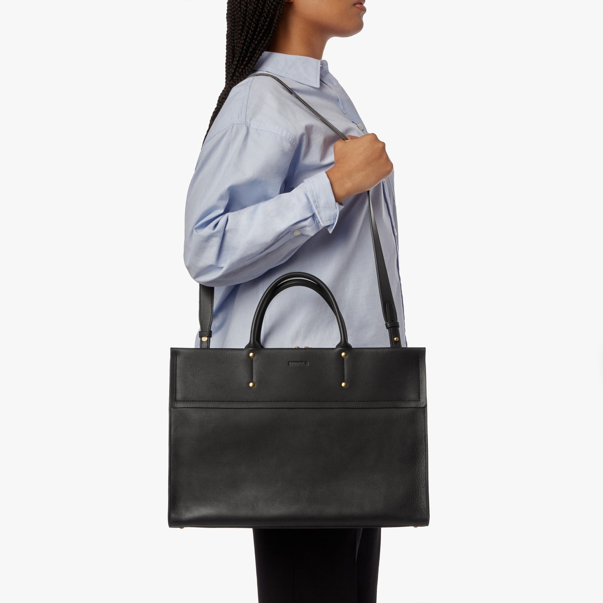 A person in a light blue button-up shirt carries the Shinola Day Satchel - Black, a large leather bag with top handles and a shoulder strap, under their arm against a plain white background.