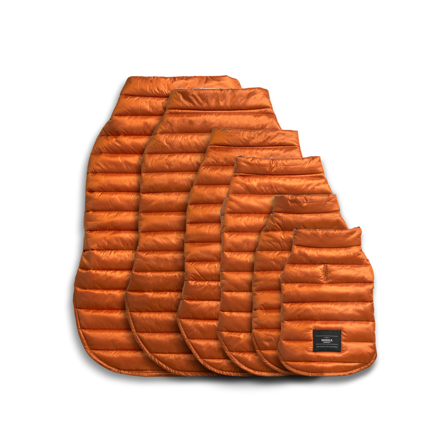 Five orange Shinola Reversible Puffer Dog Coats in various sizes are displayed overlapping on a white background. The smallest coat, resembling a cozy dog jacket, features a black patch with text near the bottom.