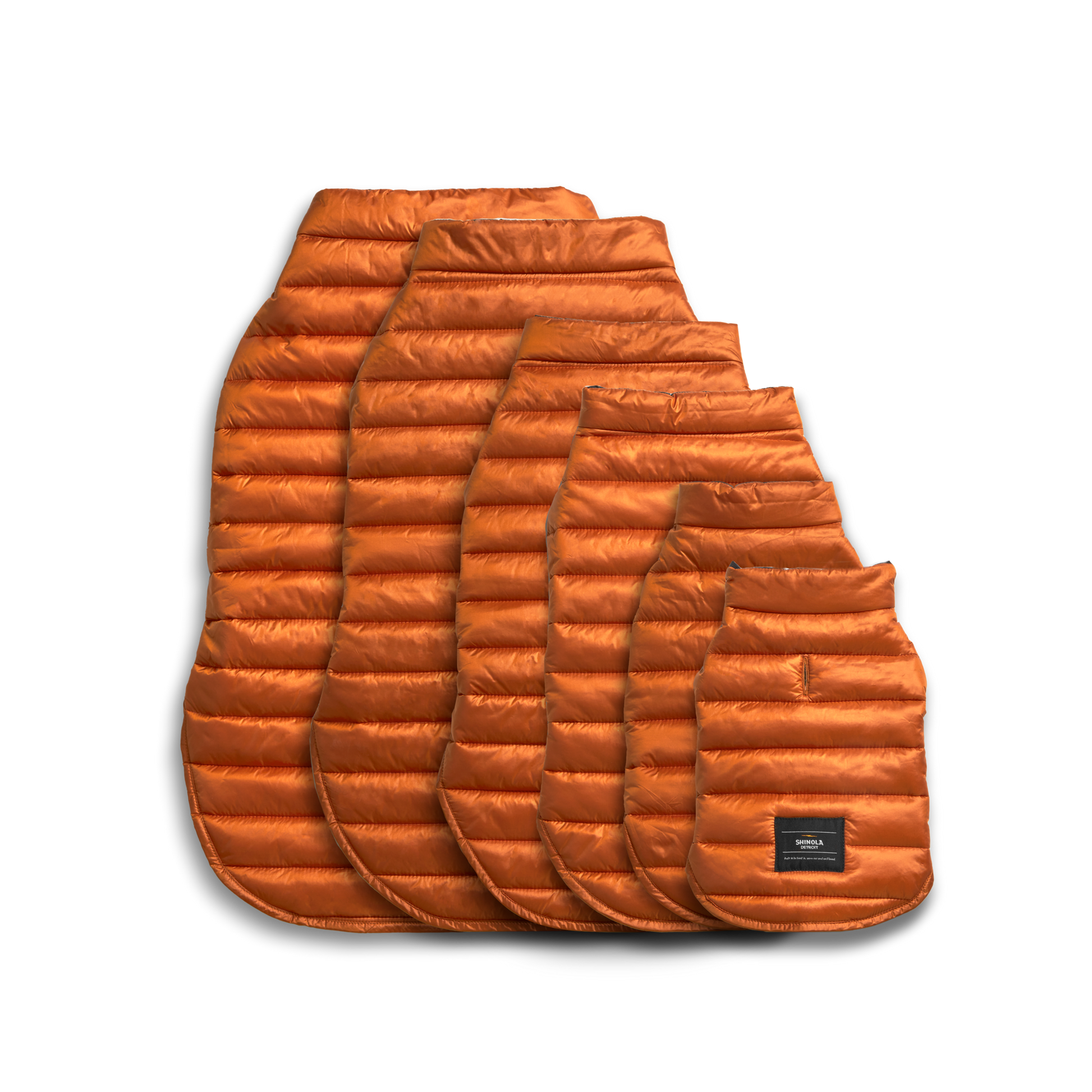 Five orange Shinola Reversible Puffer Dog Coats in various sizes are displayed overlapping on a white background. The smallest coat, resembling a cozy dog jacket, features a black patch with text near the bottom.