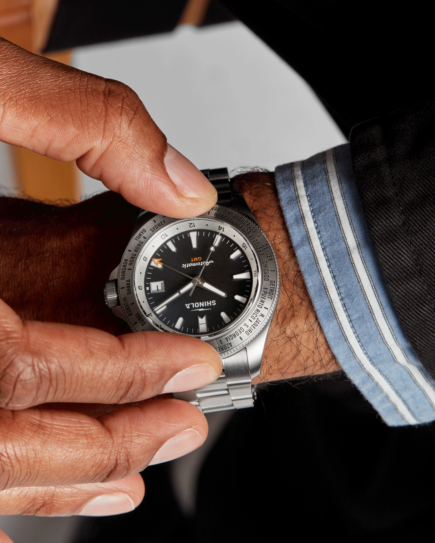 A close-up of a person adjusting a silver Shinola Monster Worldtimer GMT Watch with a world timer bezel on their left wrist, paired with a dark suit and blue-striped dress shirt.