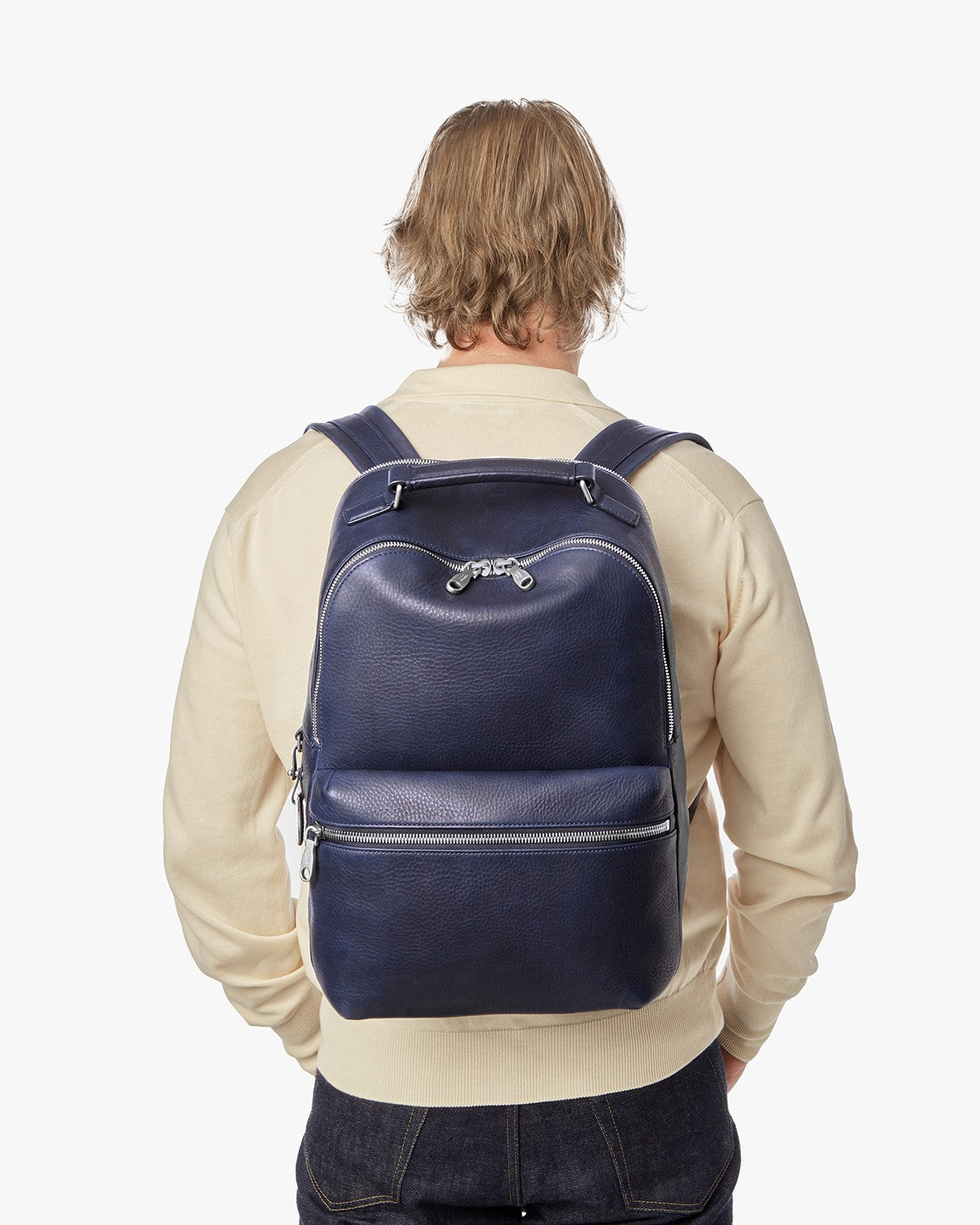 A blond-haired person in a cream sweater and dark jeans is shown from the back carrying the Shinola Portfolio Briefcase - Stone against a plain white background.