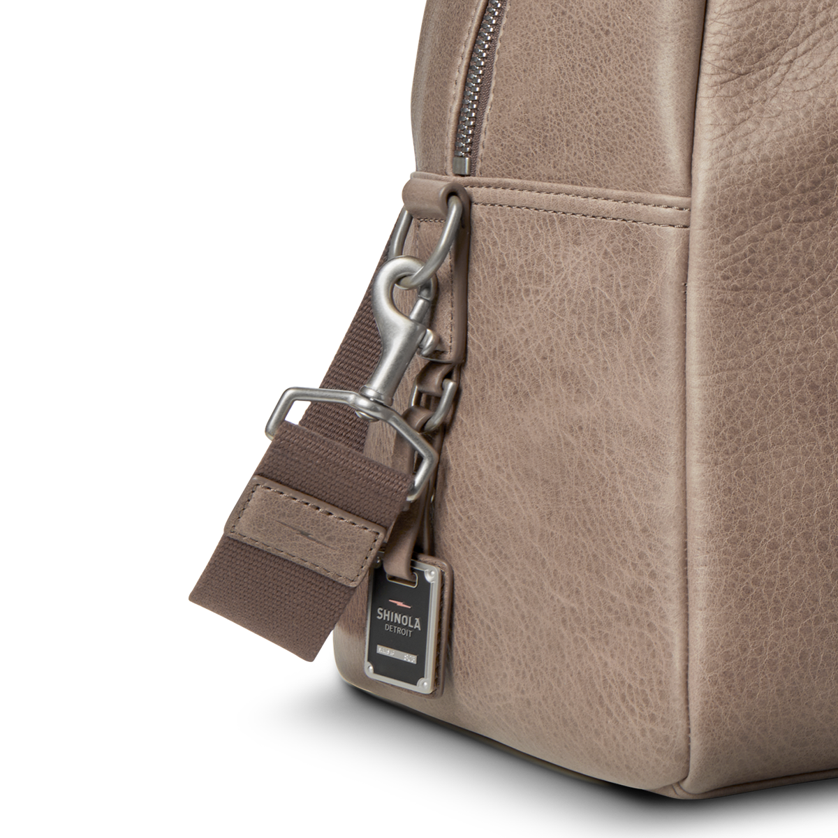 Close-up of the Shinola Runwell Duffle Bag - Stone, featuring taupe natural grain leather, a metal clip on the brown strap, and a small rectangular tag engraved with the Shinola brand name.