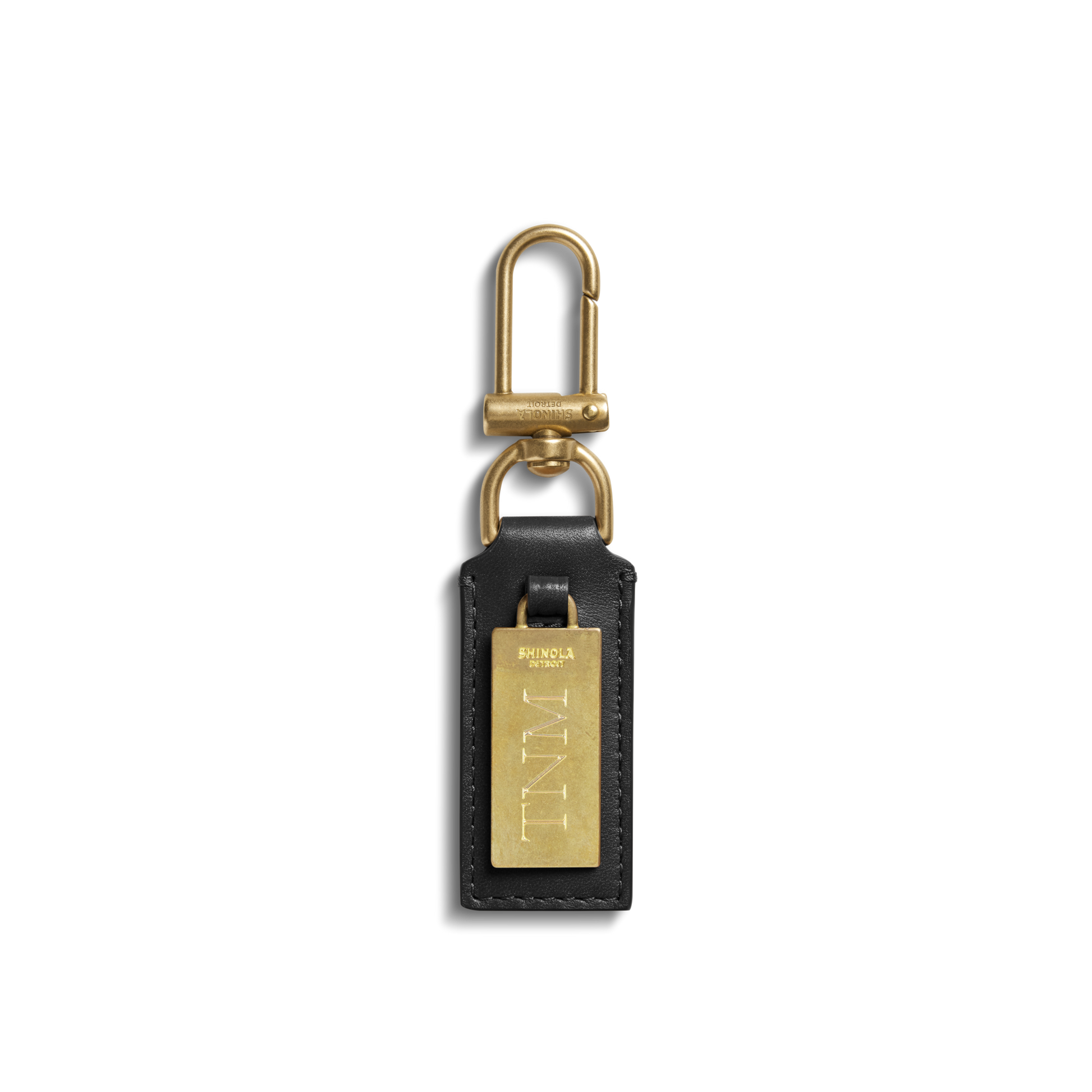 The Birdy Keychain - Black by Shinola is a natural leather key fob with a rectangular gold engravable plate engraved "TNML," attached to a brass clasp and ring, shown on a plain white background.