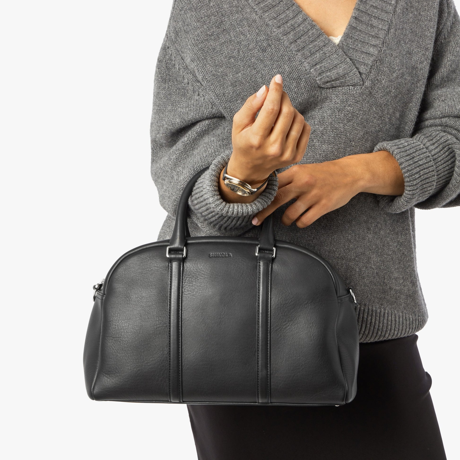 A person in a loose grey sweater and black skirt holds the Shinola Runwell Satchel - Black, a structured leather shoulder bag with two handles and vertical seams. The style recalls classic satchels. The person's face is not shown.