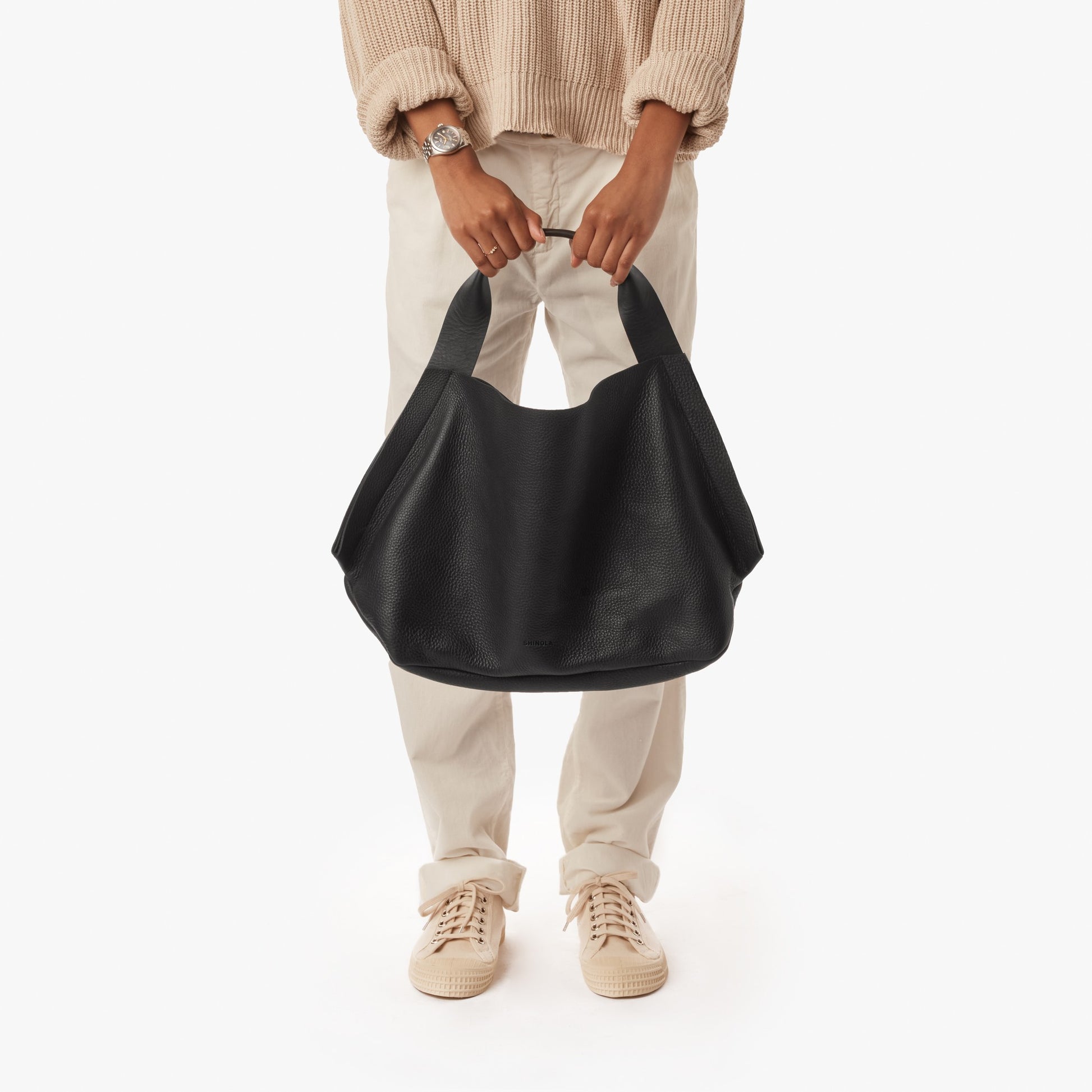 A person in a beige sweater, light pants, and beige sneakers holds the Shinola Derby Shoulder Bag - Black. The spacious black leather handbag has an open compartment and card pocket, shown against a plain white background.