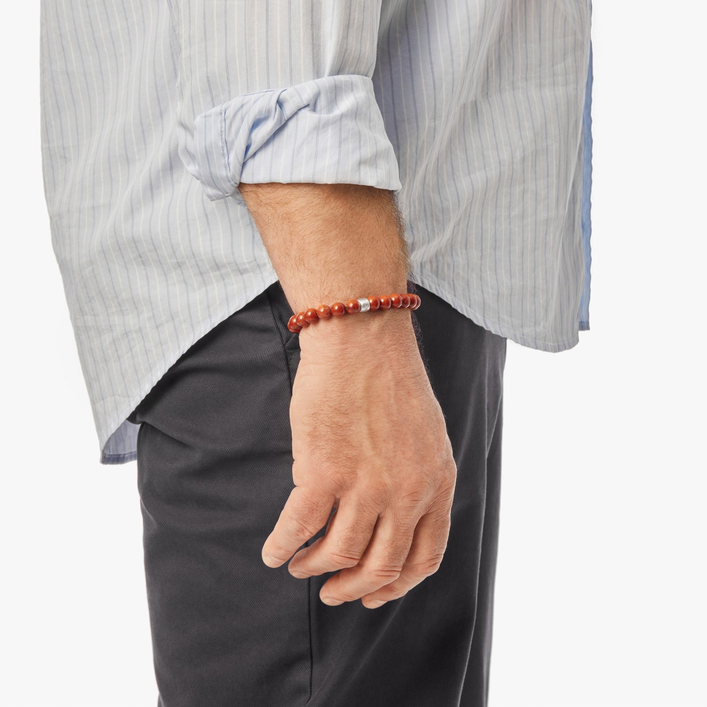 A person with their left arm relaxed displays the Shinola 8mm Beaded Red Jasper Bracelet – Red Jasper, featuring a sterling silver accent, over a light blue rolled-up striped shirt and dark pants against a white background.