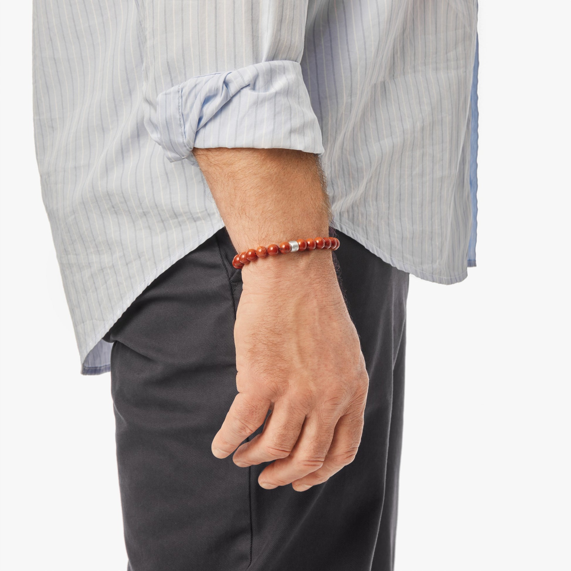 A person with their left arm relaxed displays the Shinola 8mm Beaded Red Jasper Bracelet – Red Jasper, featuring a sterling silver accent, over a light blue rolled-up striped shirt and dark pants against a white background.