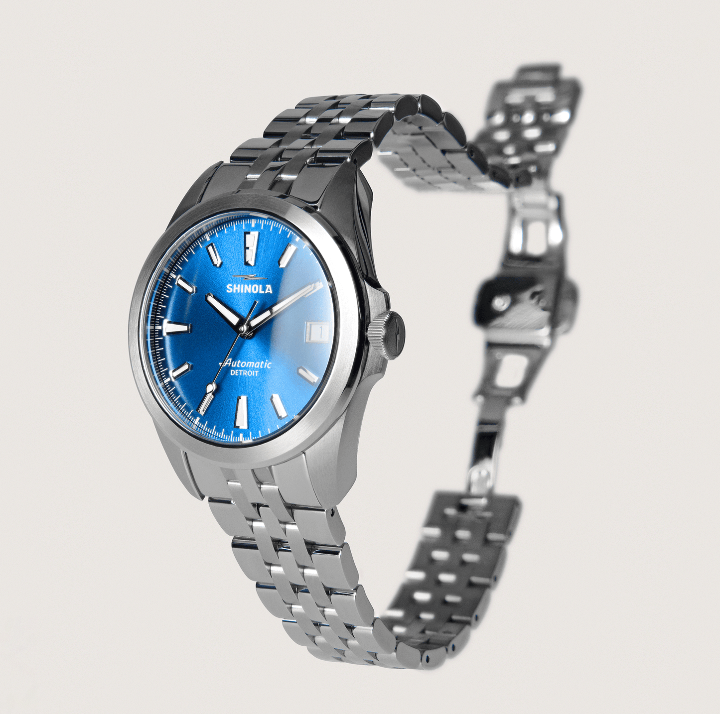 The Shinola Circadian Monster Automatic Watch - Daylight Blue features a silver metal band, blue face, silver hour markers, and date display. Its clasp is open, shown against a plain light background.