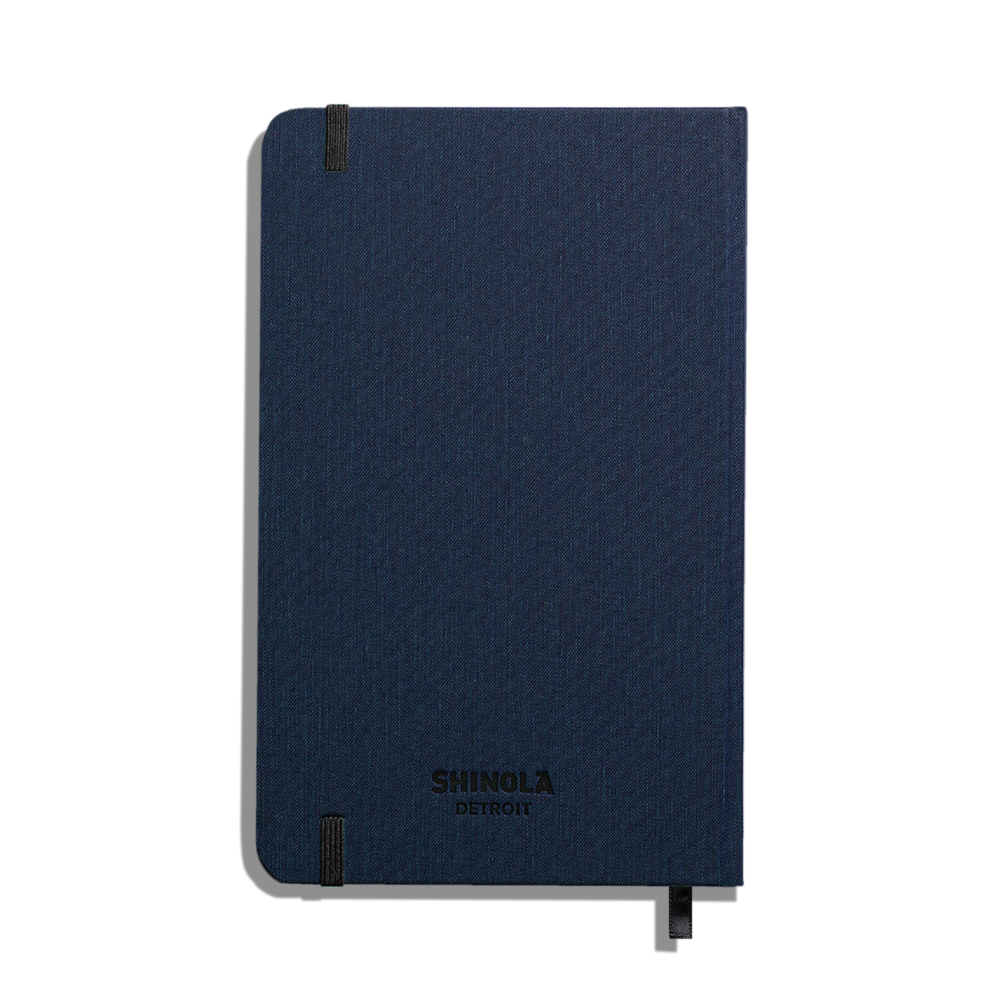 A closed Shinola Medium Hard Ruled Journal in navy with black elastic bands at two corners, smyth-sewn binding, and a matching ribbon bookmark is shown against a plain white background.
