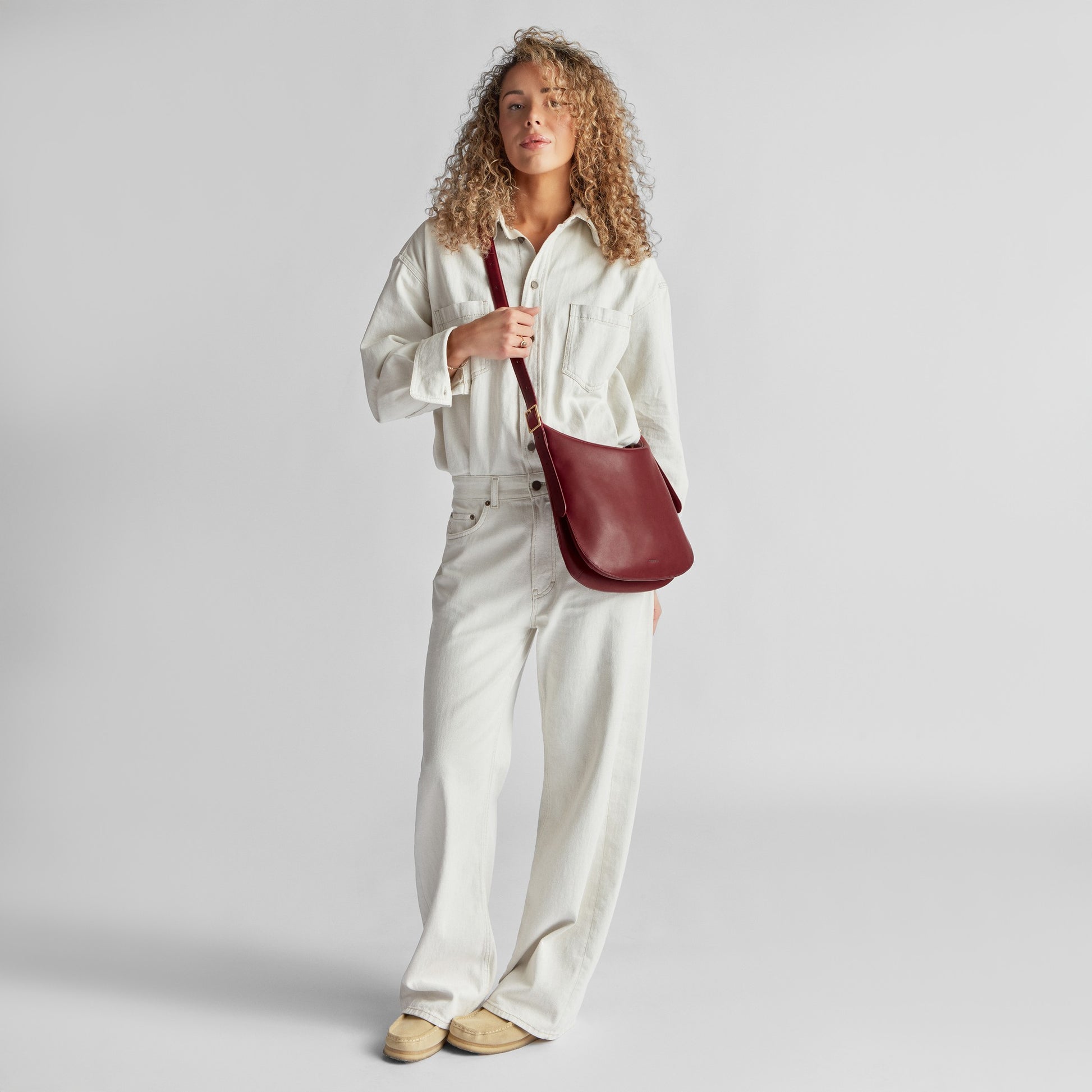 Person with curly hair wears a loose-fitting white jumpsuit, tan shoes, and carries the Shinola Medium Bixby Crossbody in Ruby—a versatile accessory—while standing against a plain light gray background.