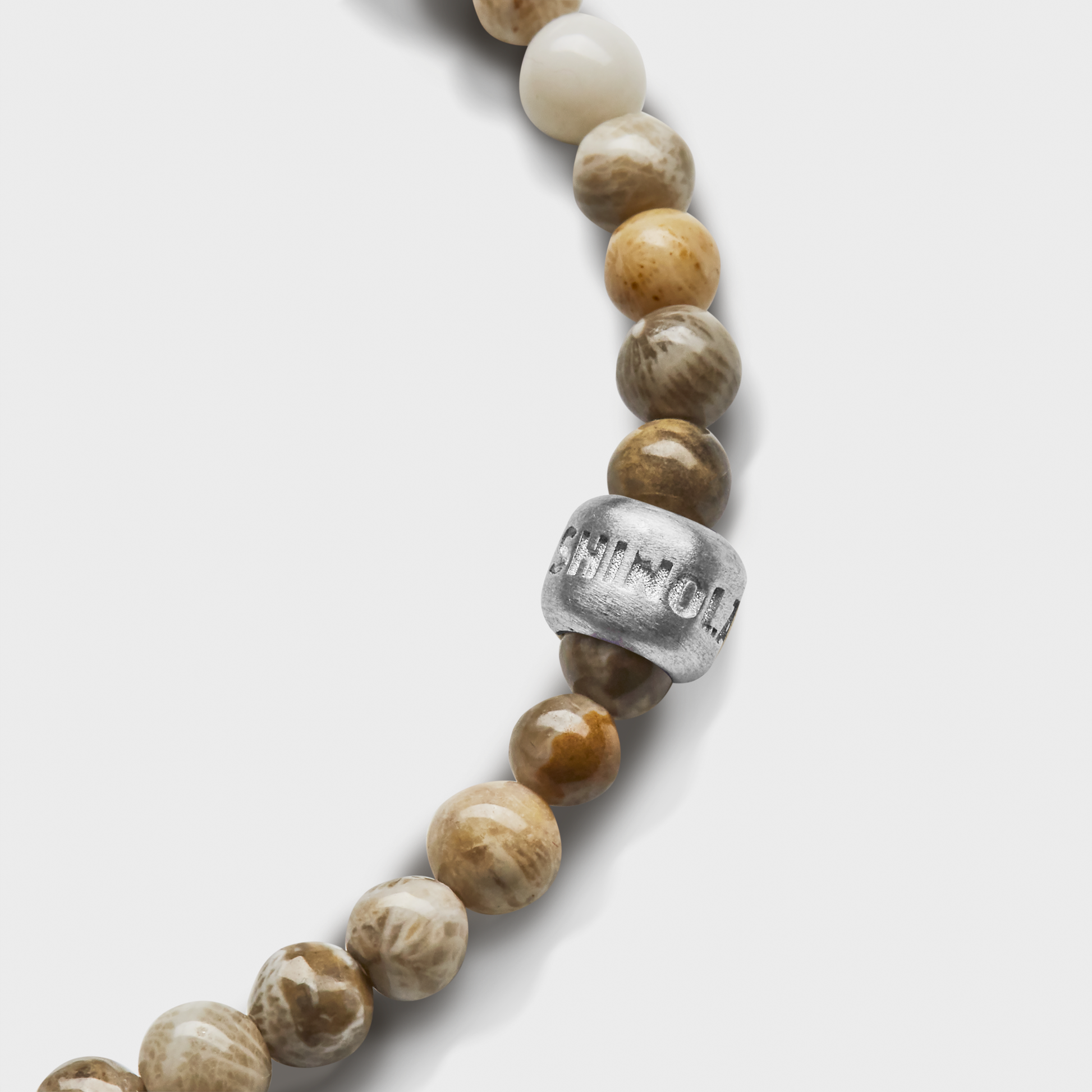 A close-up of the Shinola 4mm Beaded Petoskey Bracelet, featuring round, earth-toned polished Petoskey stones and a sterling silver accent bead engraved with letters, set against a white background.