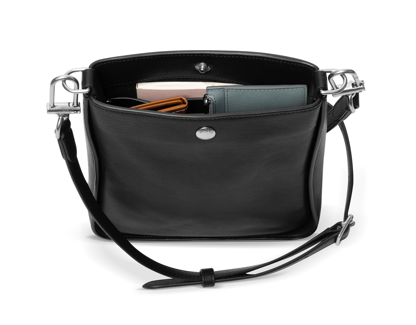 The Shinola Mini Pocket Crossbody Bag in black, shown open with a green wallet, a pink rectangular item, and an orange-striped item inside, features a snap closure and an adjustable leather strap draped in front.